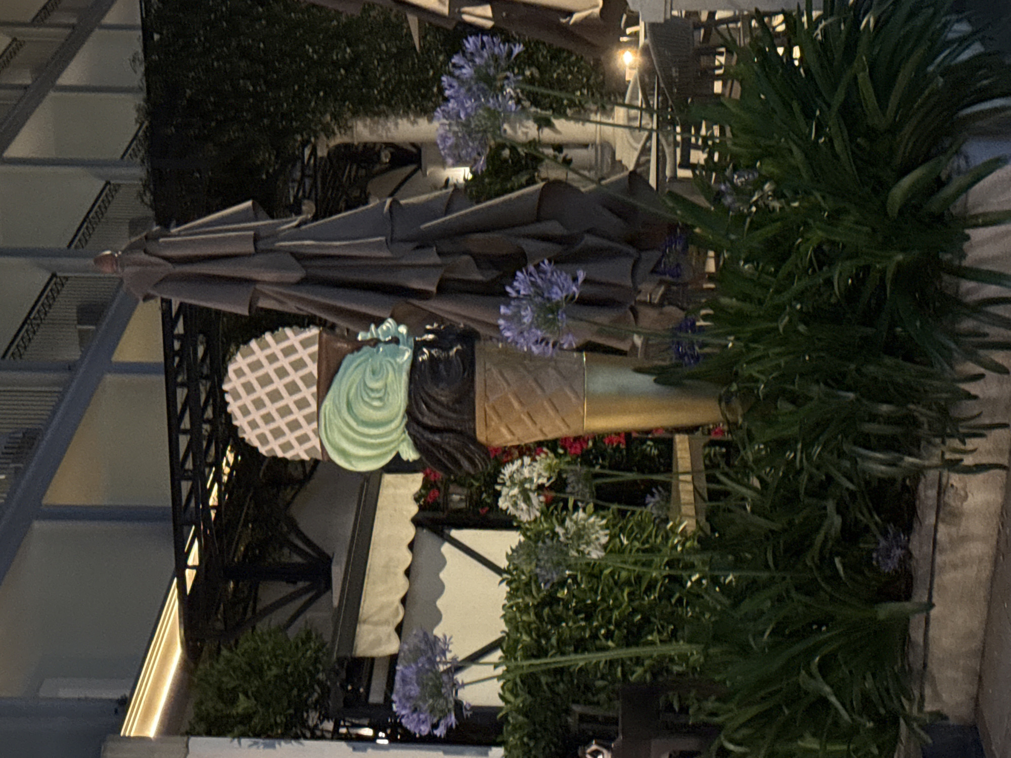 A statue of a giant ice cream cone is displayed outdoors in Budva, Montenegro. The cone is made of a tan material and topped with three scoops of ice cream in different colors: black, mint green, and white with a diamond pattern. The ice cream cone is surrounded by lush green foliage and purple and white flowers. In the background, there is a building with outdoor seating and umbrellas. The lighting suggests it is evening.