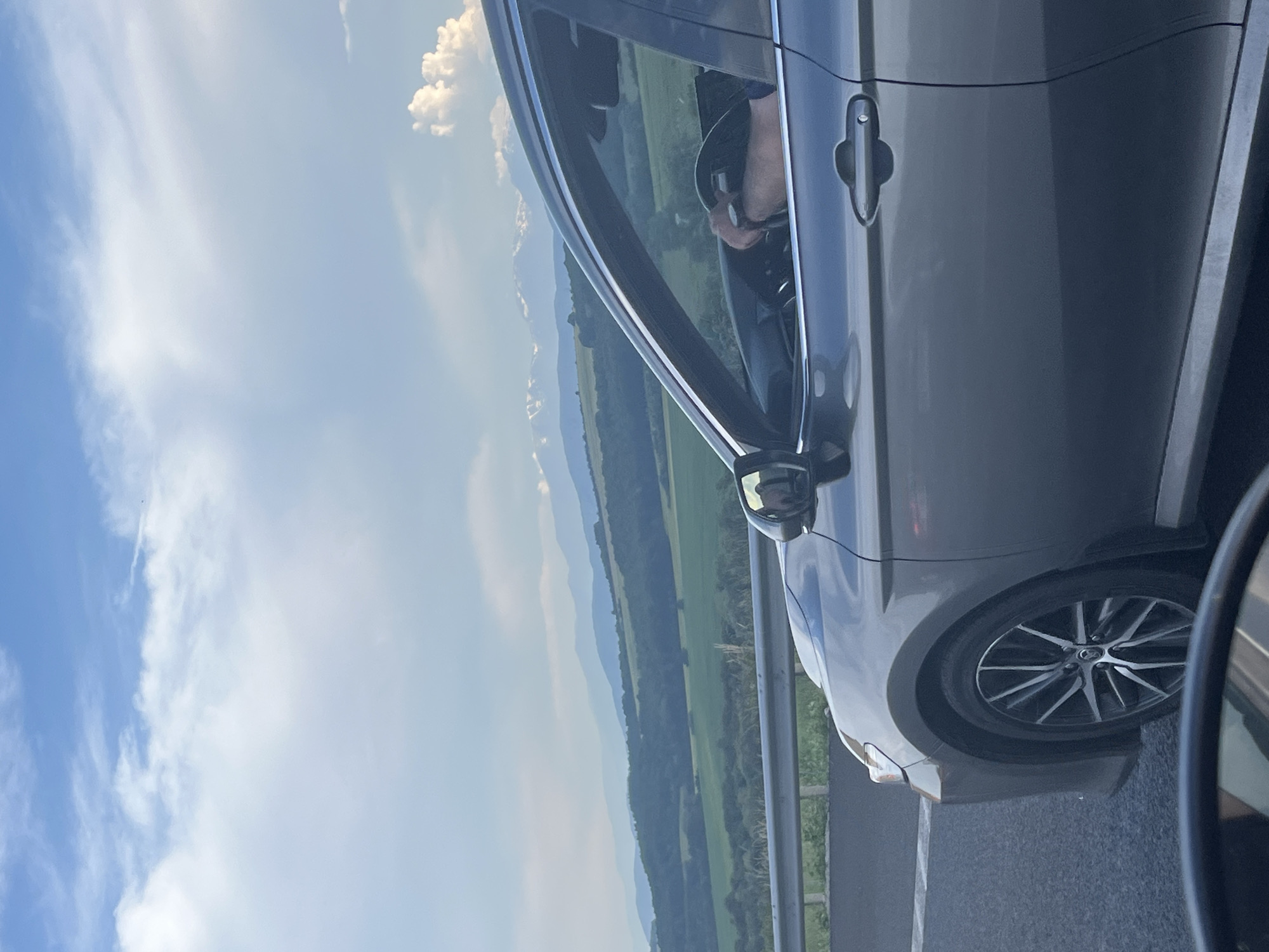 The image shows a person driving a gray car on a road, with a scenic landscape in the background. The sky is a bright blue with scattered white clouds, suggesting a clear and sunny day. The landscape features rolling green hills and distant mountains, creating a sense of depth and natural beauty. The car's interior is partially visible, showing the steering wheel and the driver's arm. The side mirror of the car also reflects the surrounding environment, offering another glimpse of the scenery. The image appears to be taken during the daytime, likely late morning or early afternoon. The overall mood is peaceful and serene, capturing a moment of travel through a picturesque rural area. The location is specified as Vakarel, Bulgaria, which adds context to the mountainous terrain and greenery. There is no visible text in the image.