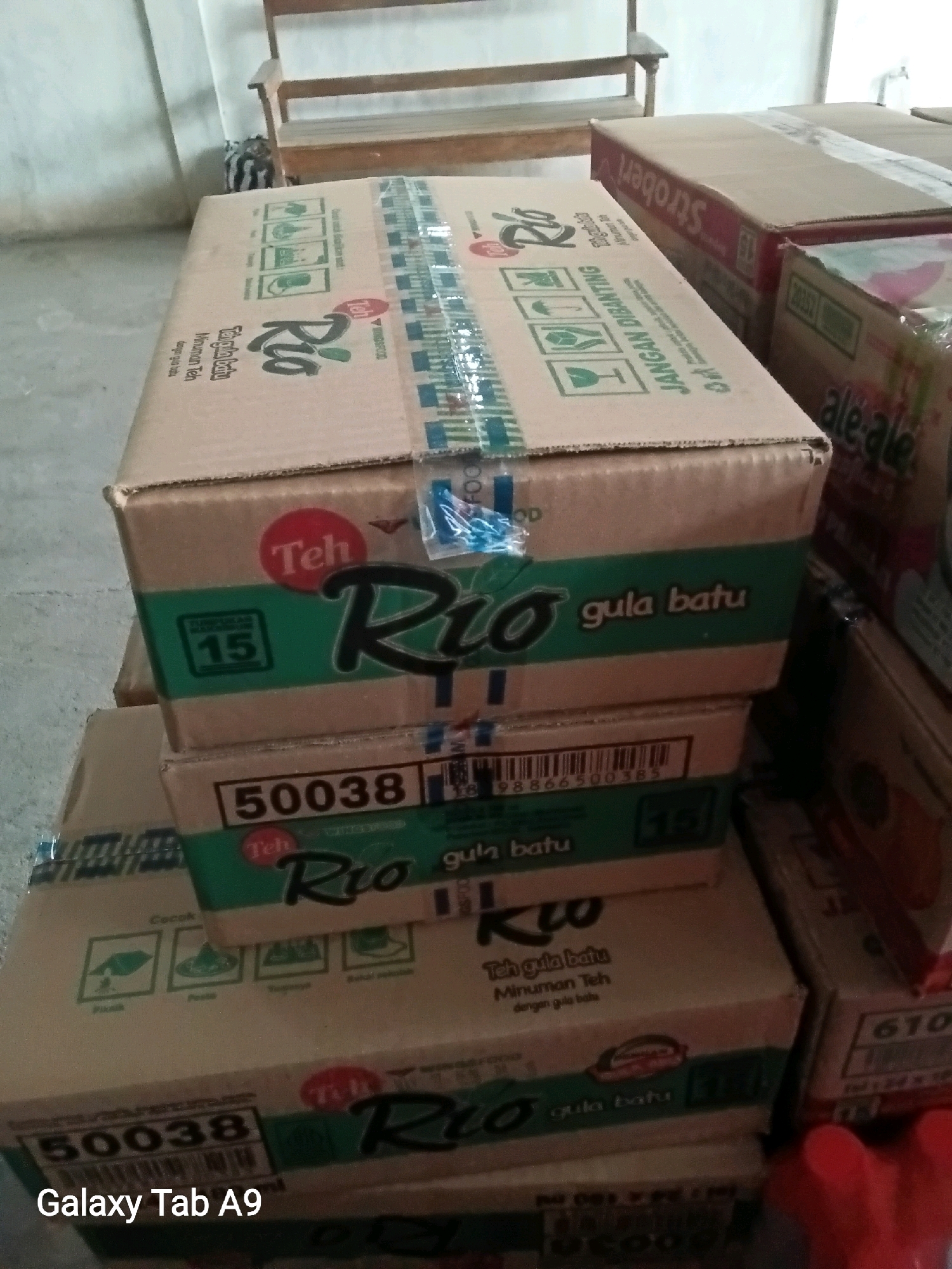 The image shows a stack of cardboard boxes on a wooden floor in an indoor environment. The boxes are of various sizes and appear to contain tea products, with labels featuring the brand "Rio" and "Teh Gula Batu" (Rock Sugar Tea). Some boxes have red circular logos with "Teh" written on them. Other visible text includes "JANGAN DIBANTING" (Do not slam), which suggests caution during handling. There are also numerical codes and barcodes on the boxes, such as "50038" and "15". In the background, a portion of a wooden bench or chair is visible. The lighting appears to be artificial, and the overall scene suggests a storage or retail environment. There are no people or animals present in the image.