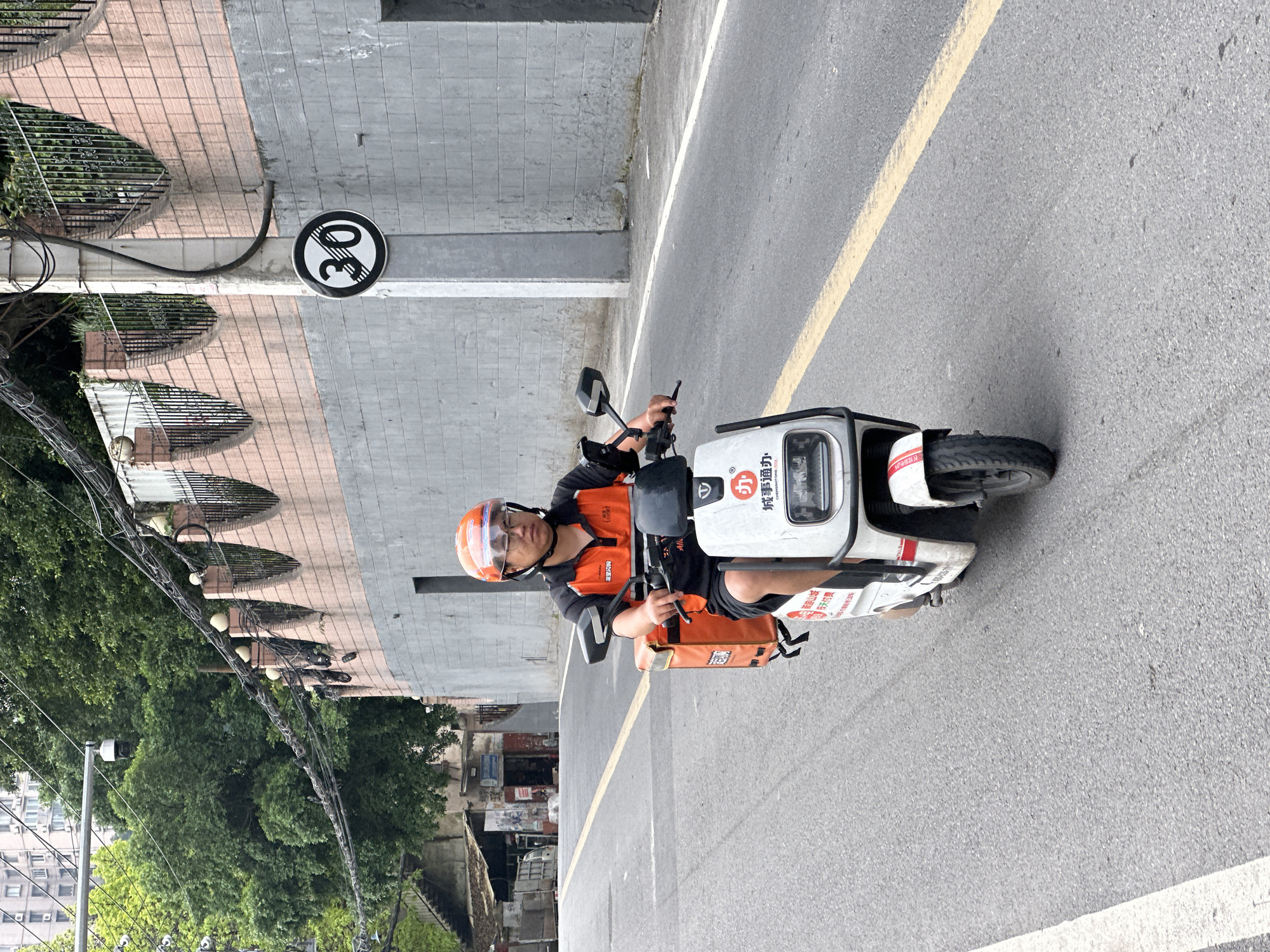 A delivery driver on a white scooter in Jiulongpo District, China. The driver wears an orange helmet and uniform. A "30" speed limit sign stands nearby, alongside a building with barred windows. Trees and power lines are visible in the background. The scooter is in the street on the right.