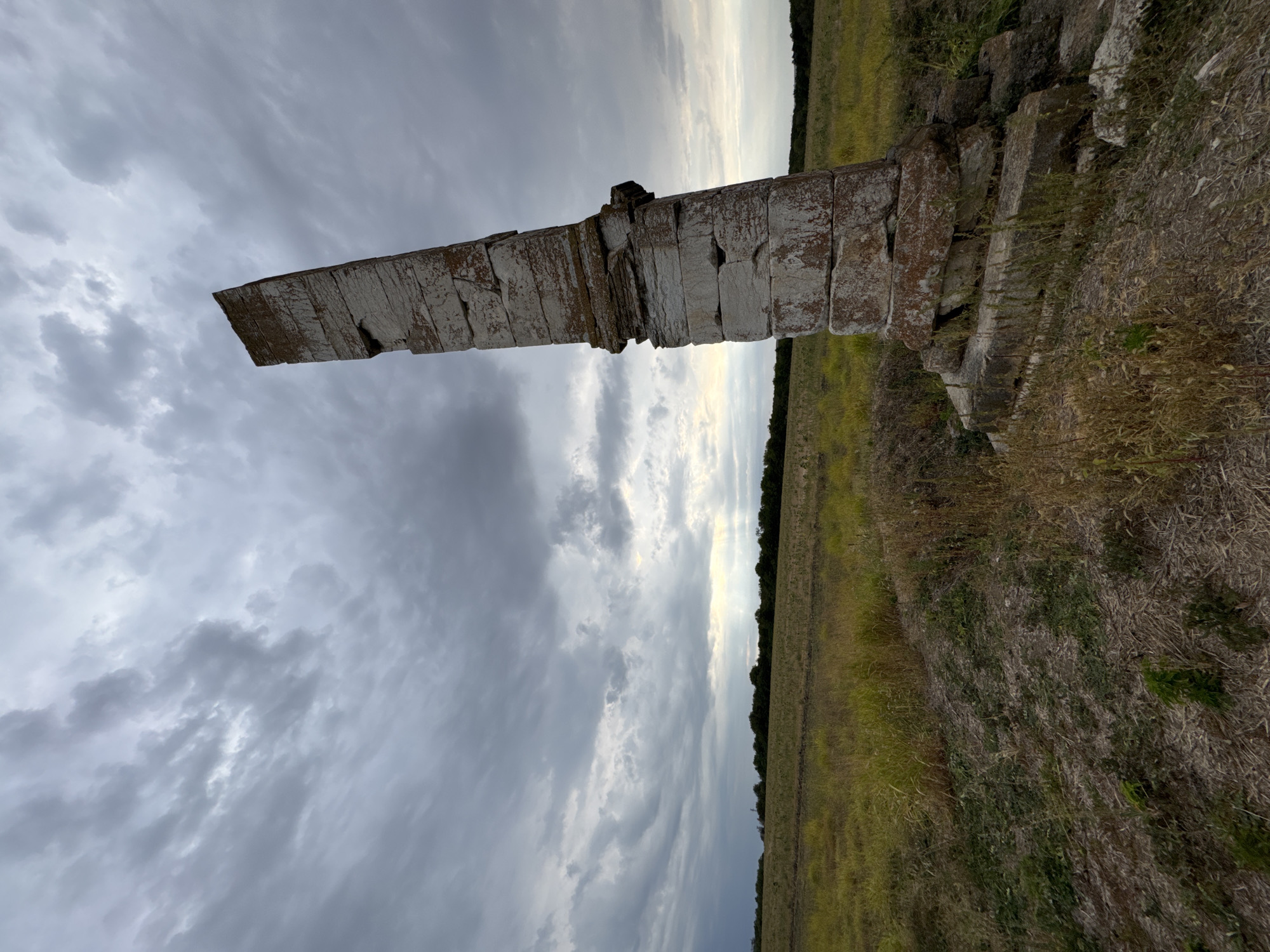 The image shows a tall, weathered stone obelisk or pillar standing in a grassy field under a cloudy sky. The obelisk is constructed from stacked rectangular stones, many of which appear chipped, cracked, and stained with reddish-brown and greenish discoloration, suggesting age and exposure to the elements. The obelisk is tilted significantly to the right.
The field surrounding the obelisk is covered in dry, yellowish-brown grass and some greener patches. In the distance, a line of trees marks the horizon. The sky is filled with dramatic, heavy gray clouds, with a sliver of lighter sky and sunlight peeking through on the right side of the frame, suggesting either dawn or dusk, or an approaching storm. The perspective is low, looking up at the obelisk and the sky, emphasizing its height and the vastness of the environment.
The location is identified as Lesicheri, Bulgaria.