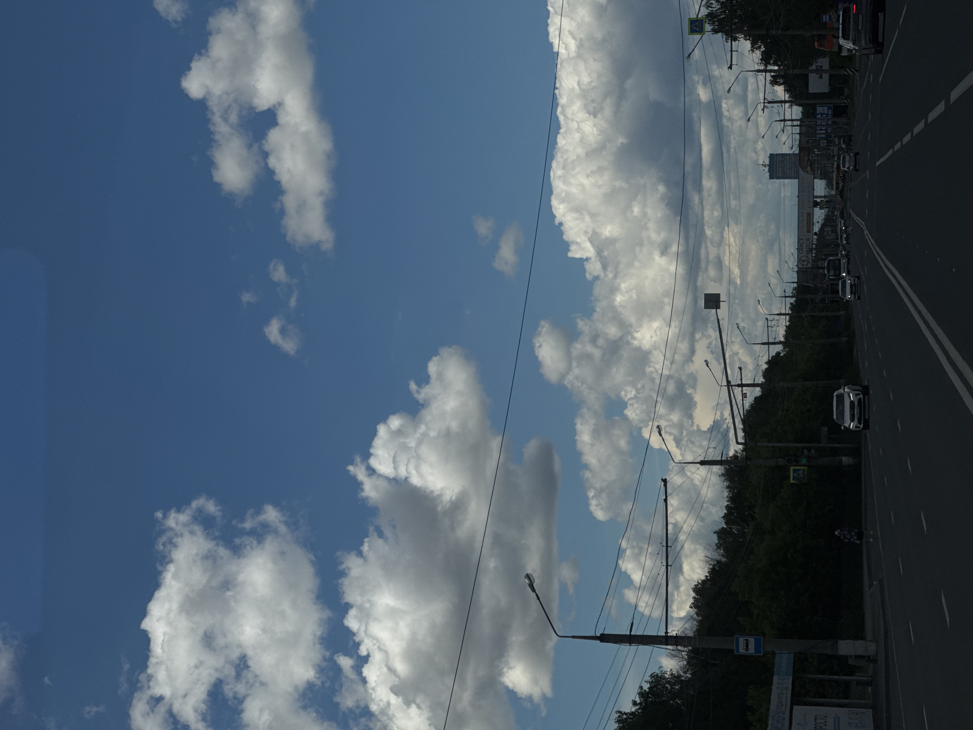 The image captures a street view in the city of Unknown, Unknown, under a partly cloudy sky. A road with double white lines stretches into the distance, lined with lampposts and power lines. There are some trees along the side of the road. Vehicles can be seen on the road. There is a bus stop on the right side of the road. The sky is blue with numerous white cumulus clouds.