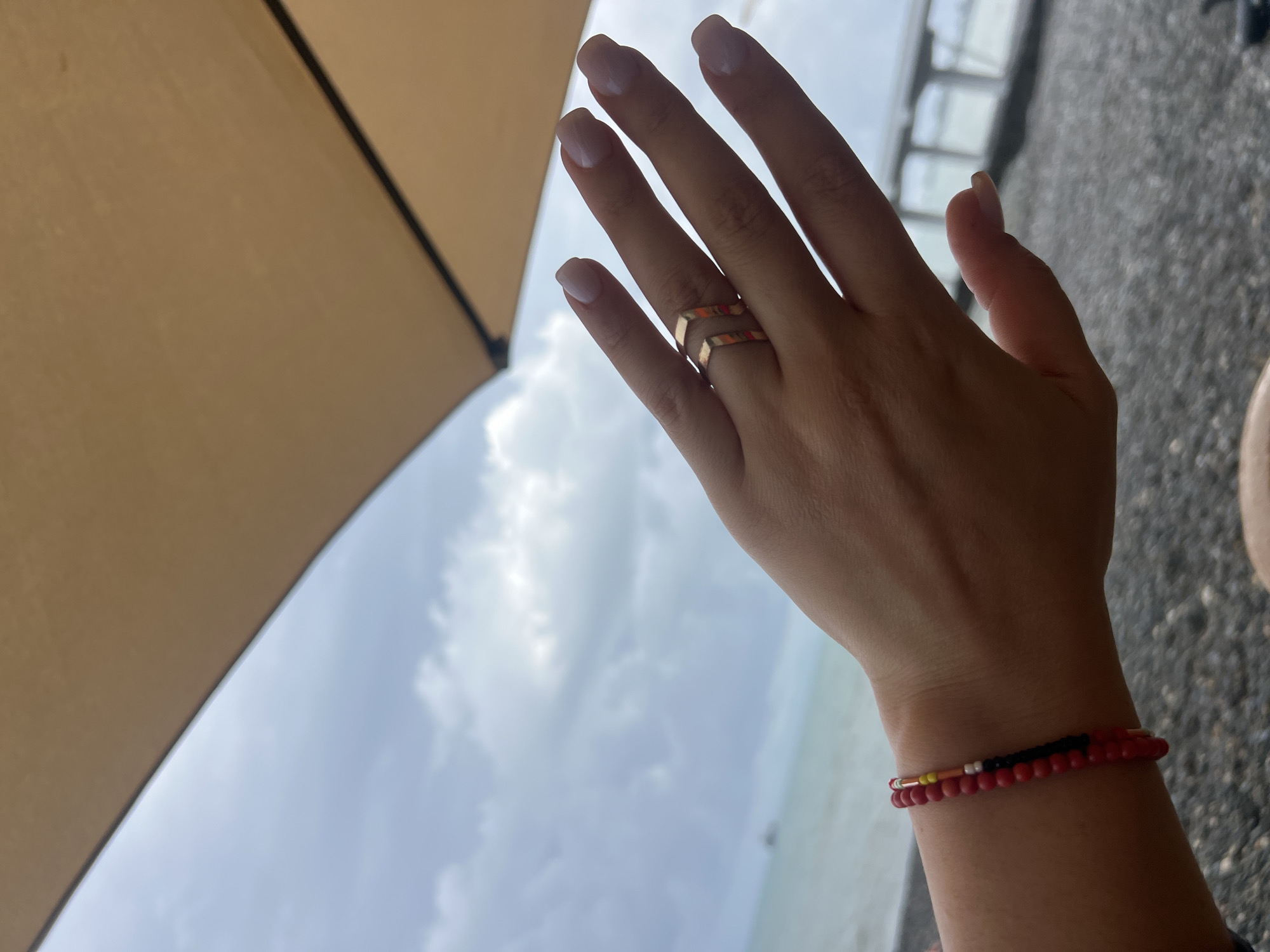A woman's hand, adorned with a rose gold chevron ring and a beaded bracelet featuring red, black, and yellow beads, is held up against a backdrop of a cloudy sky and a body of water. The hand is positioned in the foreground, with the fingers slightly splayed. The background suggests a beach or coastal setting, with blurred details of a structure that could be a pier or boardwalk. The overall lighting is overcast, indicating a cloudy day. The location is identified as Sokhumi, Abkhazia.