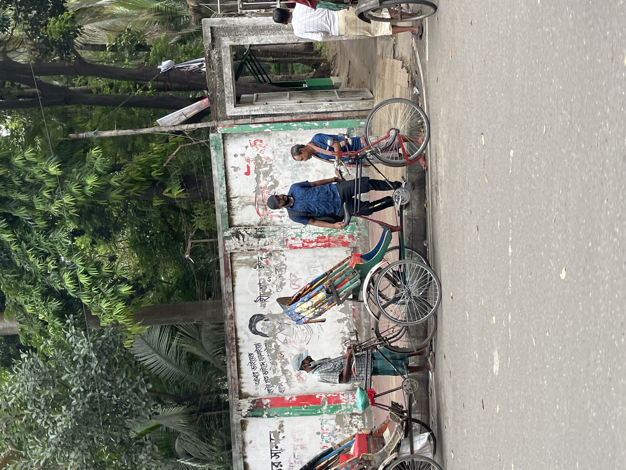 This image captures a vibrant street scene in Dhaka, Bangladesh, featuring several men, traditional rickshaws, and lush greenery alongside a concrete wall. The scene is set during the daytime under diffused lighting, indicating either an overcast sky or late morning/early afternoon.
On the left side of the frame, dense foliage and trees create a natural backdrop. A weathered concrete wall runs horizontally across the middle, separating the greenery from a paved street or pathway on the right. This wall is a focal point, adorned with peeling paint in red, green, and white stripes, colors reminiscent of the Bangladeshi flag. It also features a black-and-white mural of a man, commonly identifiable as Bangabandhu Sheikh Mujibur Rahman, along with various inscriptions in Bengali script.
Several individuals are present, predominantly men, engaged with bicycles and traditional pedal-powered rickshaws. In the foreground, a man wearing a dark blue patterned shirt, black pants, and a baseball cap stands next to a bicycle. Adjacent to him, another man in a blue shirt appears to be holding onto a rickshaw. Further along the wall, a third man with a light-colored cap and a checkered shirt is seen attending to a rickshaw with a decorated passenger seat. Near the upper right, by a concrete doorway in the wall, another man in a white and striped shirt and a lungi stands, looking towards the street. The legs of another person are partially visible at the bottom right.
The overall activity suggests a typical urban setting where people and rickshaws are part of the daily commute or waiting for passengers. A bamboo pole with attached objects, possibly lights or signs, extends from a structure behind the wall, adding to the bustling street atmosphere.