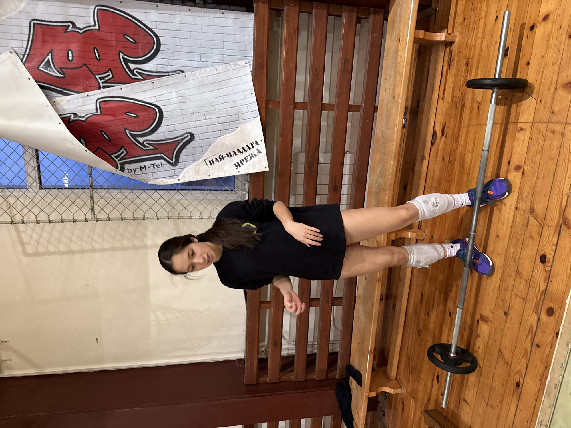 The photograph shows a woman standing next to a barbell in what appears to be a gymnasium. The woman has long dark hair and is wearing a black t-shirt, knee pads, and purple and blue shoes. The barbell has weights on each end. The background includes a wooden structure, a wall with graffiti-style art with the words "HAH," and a chain-link fence. A portion of the banner is written in Bulgarian ( "НАЙ-МЛАДАТА. МРЕЖА by M-Tel"). This scene likely occurs in Sofia, Bulgaria, as indicated by the Bulgarian language on the banner.