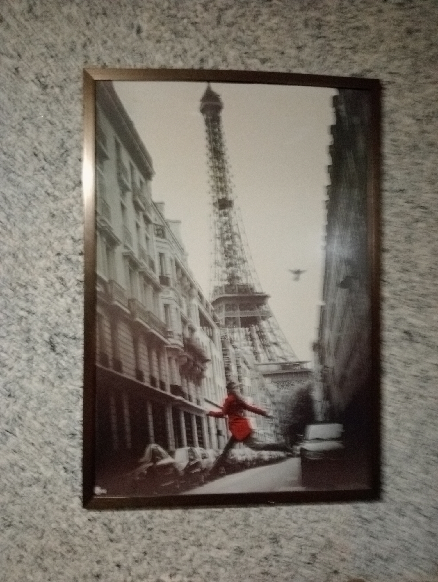A framed black and white photograph shows a street scene in Paris, France, with the Eiffel Tower prominently in the background. A woman in a red coat is captured in mid-stride, with her arms outstretched. The street is lined with multi-story buildings and parked cars. A bird is visible in flight above the street. The image has a blurred, artistic quality.