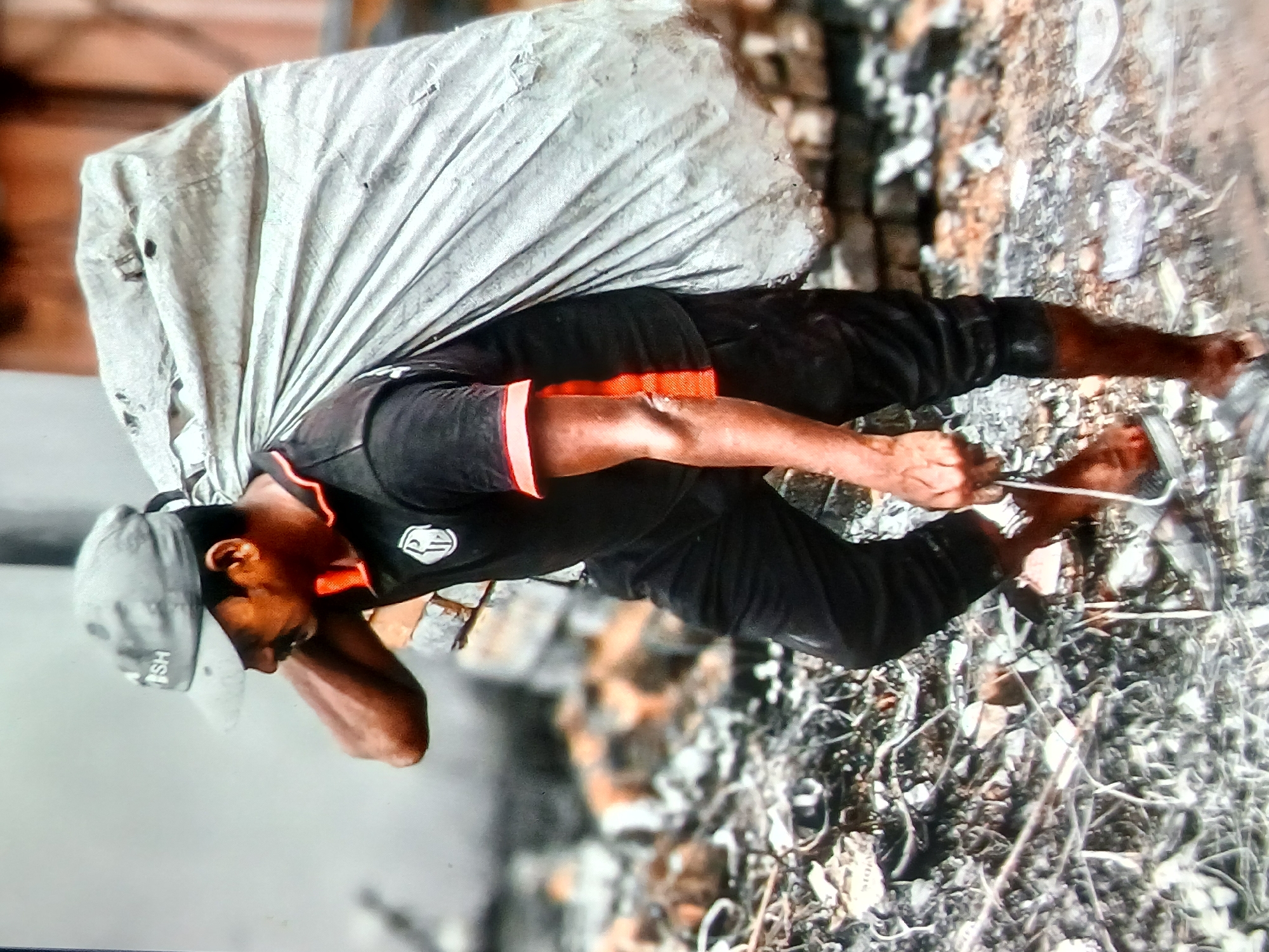 In the city of Setia Bakti, Indonesia, a man wearing a grey cap and a black t-shirt with orange accents carries a large, grey sack over his shoulder. The sack appears to be made of a tarp-like material and is filled with unknown contents. The man is walking on a debris-strewn surface, which includes broken bricks and various organic materials. His right leg is bent, and he is wearing dark pants and appears to be barefoot or wearing very minimal footwear. The background is blurred, suggesting the focus is on the man and his burden.