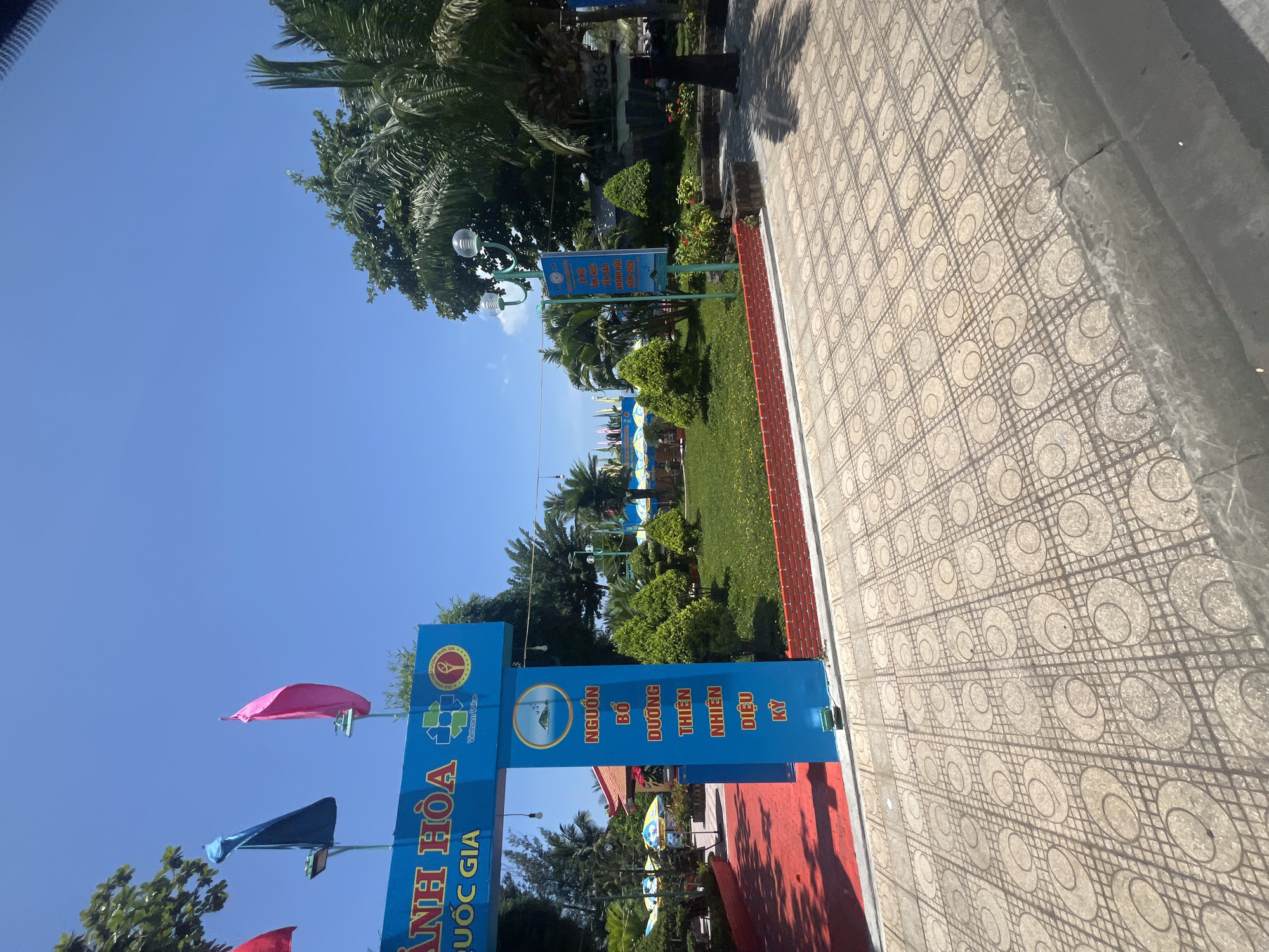 The image shows an entrance to a park or public space in Nha Trang, Vietnam. The entrance archway is painted blue with red lettering. Flags of red, blue, and pink are displayed above the arch. A blue post with Vietnamese text and a circular emblem featuring an island and birds is also visible. Beyond the entrance, a well-maintained green lawn is dotted with small, manicured bushes. In the background, there are palm trees, other lush vegetation, and umbrellas, suggesting a tropical environment. The ground in the foreground is paved with patterned tiles. A street light is attached to a green pole on the right side of the image, and a person wearing dark clothing is standing near it. The sky is clear and blue.