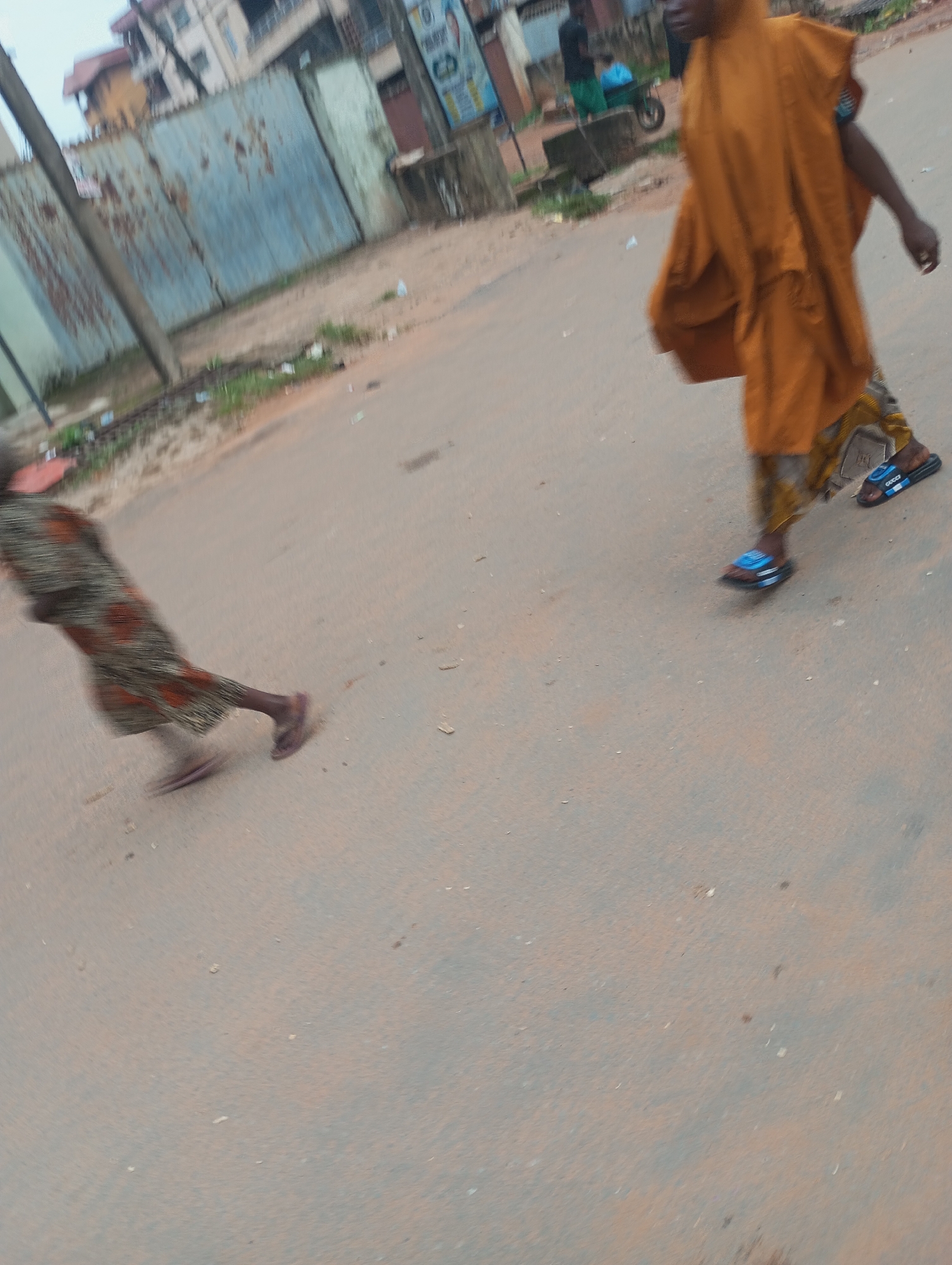 Two individuals are depicted on a paved road in Onitsha, Nigeria. The person on the right, facing left, is wearing an orange garment that appears to be an abaya or similar traditional attire, with patterned leggings and blue and black sandals. The person on the left, facing right and slightly blurred due to motion, is wearing a patterned dress and dark sandals. The background shows a corrugated metal fence, buildings, and a utility pole. The scene suggests an outdoor, urban environment.