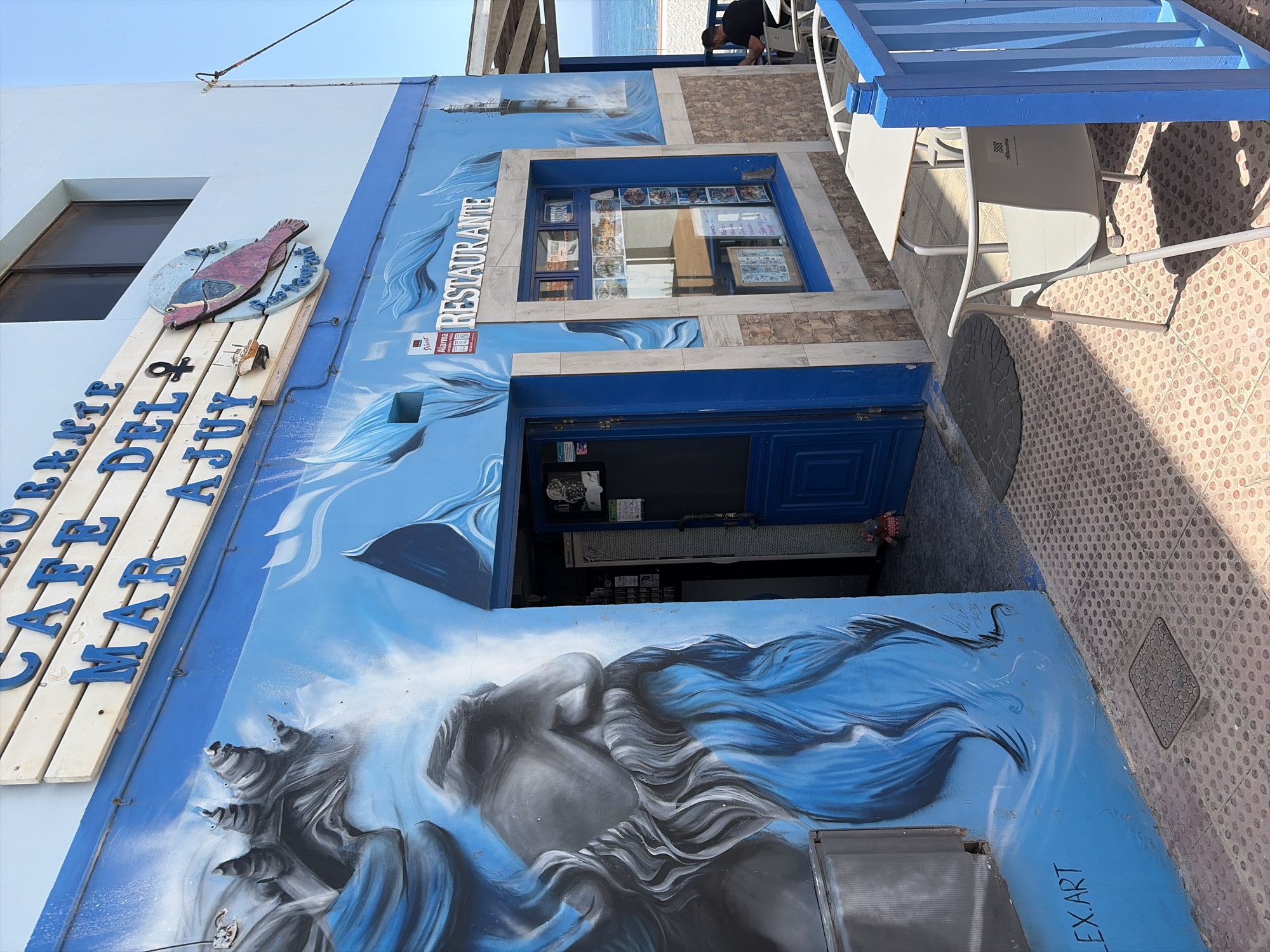 A vibrant, art-adorned cafe or restaurant building stands out with a predominantly blue and white facade. The exterior is covered in elaborate murals featuring crashing waves, a stylized lighthouse, and a large depiction of a mythical figure, possibly a sea goddess or siren, with flowing blue hair or drapery.
Outdoor seating with white tables and chairs is arranged on a tiled patio, illuminated by bright sunlight, casting distinct shadows. In the upper background, beyond the building and seating area, a sandy beach and the blue sea are visible, with a person standing near the water.
A prominent wooden sign on the left side of the building reads "CAFE DEL MAR AQUI" (Cafe of the Sea Here), with blue letters affixed to light wooden planks. Above this sign, a red-hued fish is mounted on a circular base. Another sign, above a display window, spells out "RESTAURANTE" in white letters. The display window itself showcases various food items.
Notable details include an alarm sticker with "Alarma Grimalt" and a phone number next to the "RESTAURANTE" sign. The signature "ALEX.ART" is visible on the lower right portion of the mural, indicating the artist. The sunny weather and clear blue sky suggest it's daytime, possibly midday or early afternoon. The overall scene conveys a cheerful, seaside atmosphere in Pájara, Spain.