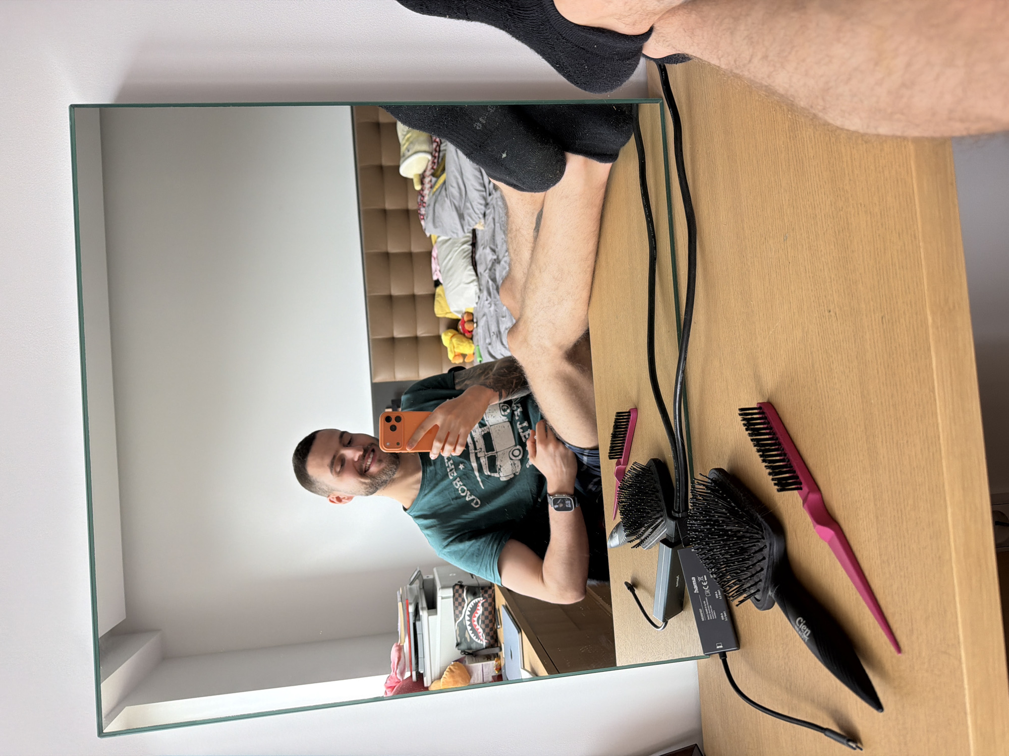 A man takes a mirror selfie in an apartment in Sofia, Bulgaria. The man, with short hair and a beard, is wearing a green t-shirt and a smartwatch, holding an orange smartphone. The mirror reflects a cluttered room with a bed, luggage, and other items. On the table in front of the mirror, there are two hairbrushes and a comb. A pair of black socks are visible in the upper part of the mirror's reflection. A charging cable lies nearby. The surrounding decor suggests a contemporary interior, with clean white walls.