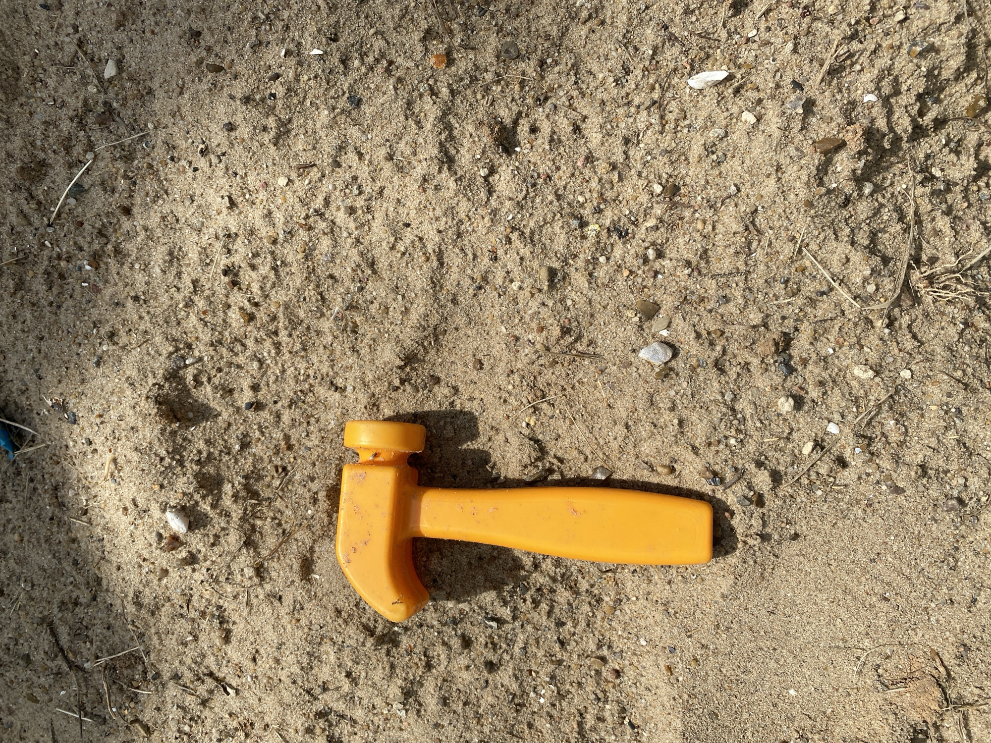 An orange toy hammer lies on a surface of coarse sand and small pebbles, casting a shadow to its right. The sand is unevenly distributed, with some areas appearing more compacted than others. Several small pieces of debris, including dried grass and twigs, are scattered across the sand. The lighting suggests it is daytime, with sunlight creating distinct shadows. No people are visible in the frame.