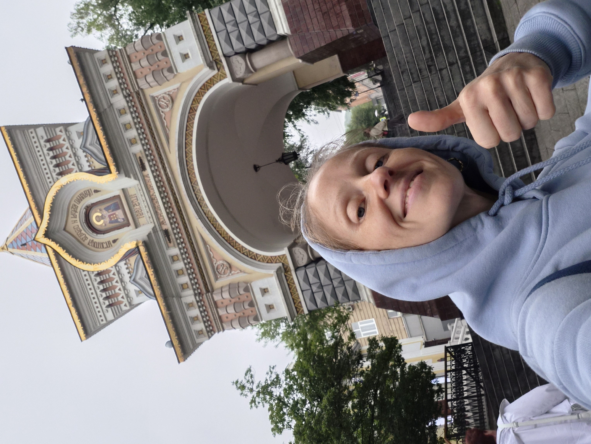 A woman wearing a light blue hooded sweatshirt smiles and gives a thumbs-up while standing in front of the Triumphal Arch in Vladivostok, Russia. The arch is decorated with intricate patterns and a depiction of a religious figure. The woman's face is close to the camera, and the arch is visible behind her. There are trees and buildings in the background, as well as a set of stairs. Another person, wearing a white hooded jacket, is visible walking up the stairs in the lower left of the image. The sky is overcast.