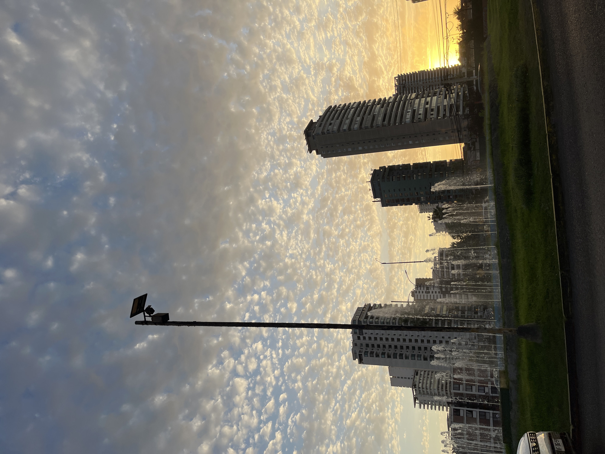 The image depicts a cityscape at sunset, possibly of Unknown, Unknown. The sky is filled with altocumulus clouds, catching the golden light of the setting sun. Buildings of varying heights dominate the midground, including several high-rise residential towers. Fountains are visible in front of the buildings, their streams of water reflecting the sunset. A lamp post is in the foreground. A road is on the right, beside a strip of green grass.