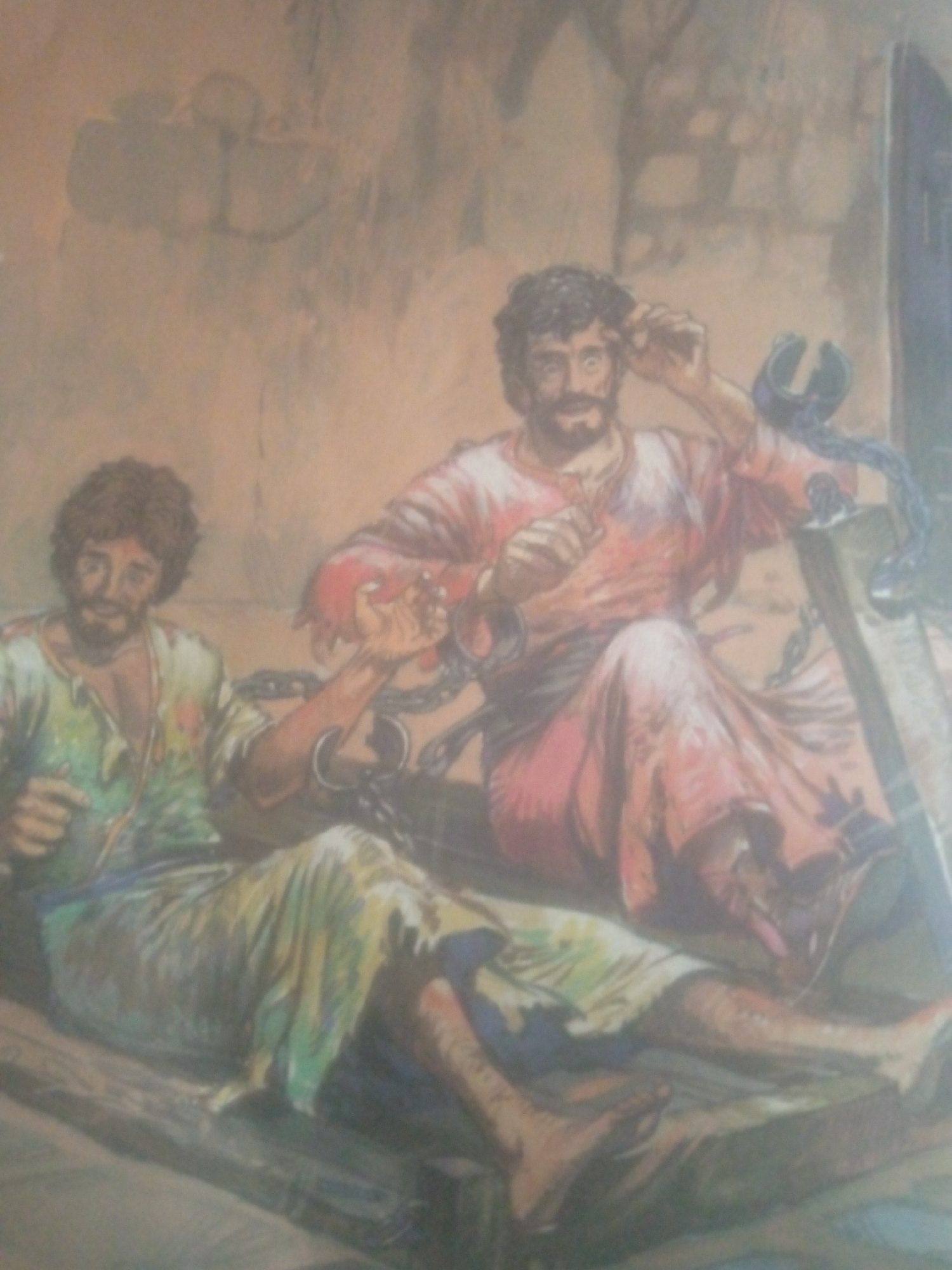 This image depicts two men in what appears to be a prison or holding cell, indicated by the rough, textured walls and the presence of chains. Both men are seated, and their attire suggests a historical or biblical context.
The man on the left, with curly brown hair and a beard, wears a tattered green and white garment. He is looking towards the viewer with a somber expression and his hands are resting on his lap. He appears to be shackled.
The man on the right, also with dark, curly hair and a beard, is dressed in a red robe. He is looking up and to the side, with one hand raised towards his head. He is also bound by chains, which are visible around his neck and wrists. These chains are also attached to some sort of structural element behind him. The overall atmosphere is one of confinement and distress. There is no visible text in the image.