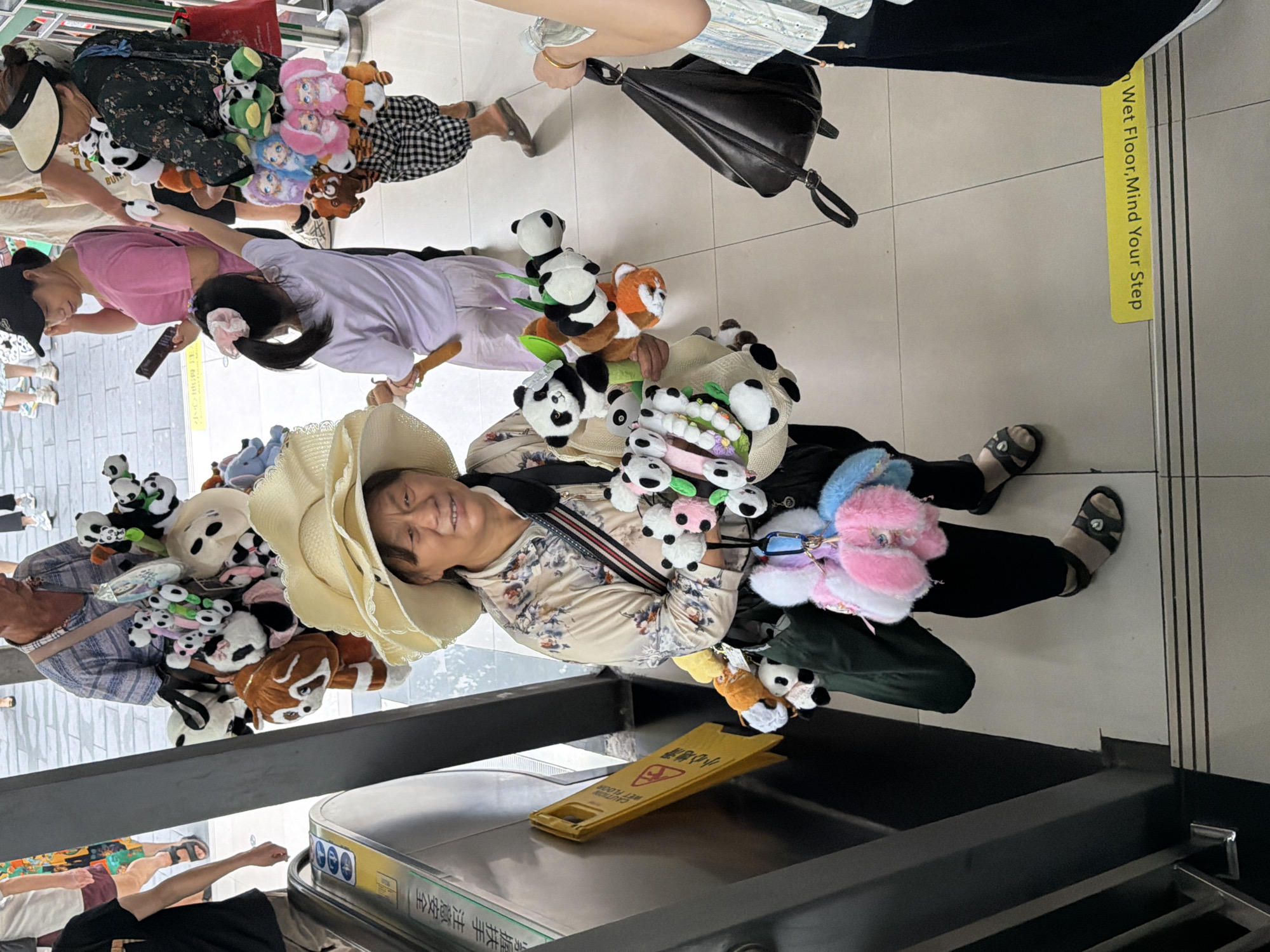 An adult East Asian woman, wearing a wide-brimmed straw hat and a floral print shirt, stands on a tiled floor next to an escalator in the Jiulongpo District, China. She is carrying an assortment of stuffed animals, primarily pandas and some colorful plush figures, suspended from her arms and a bag. The woman appears to be a vendor, as suggested by the quantity and variety of merchandise. Behind her, other people are visible, including a child reaching out towards the stuffed animals and another woman holding a phone. A yellow "Wet Floor, Mind Your Step" sign is propped against the escalator. The scene suggests a public area, possibly a shopping center or transportation hub.