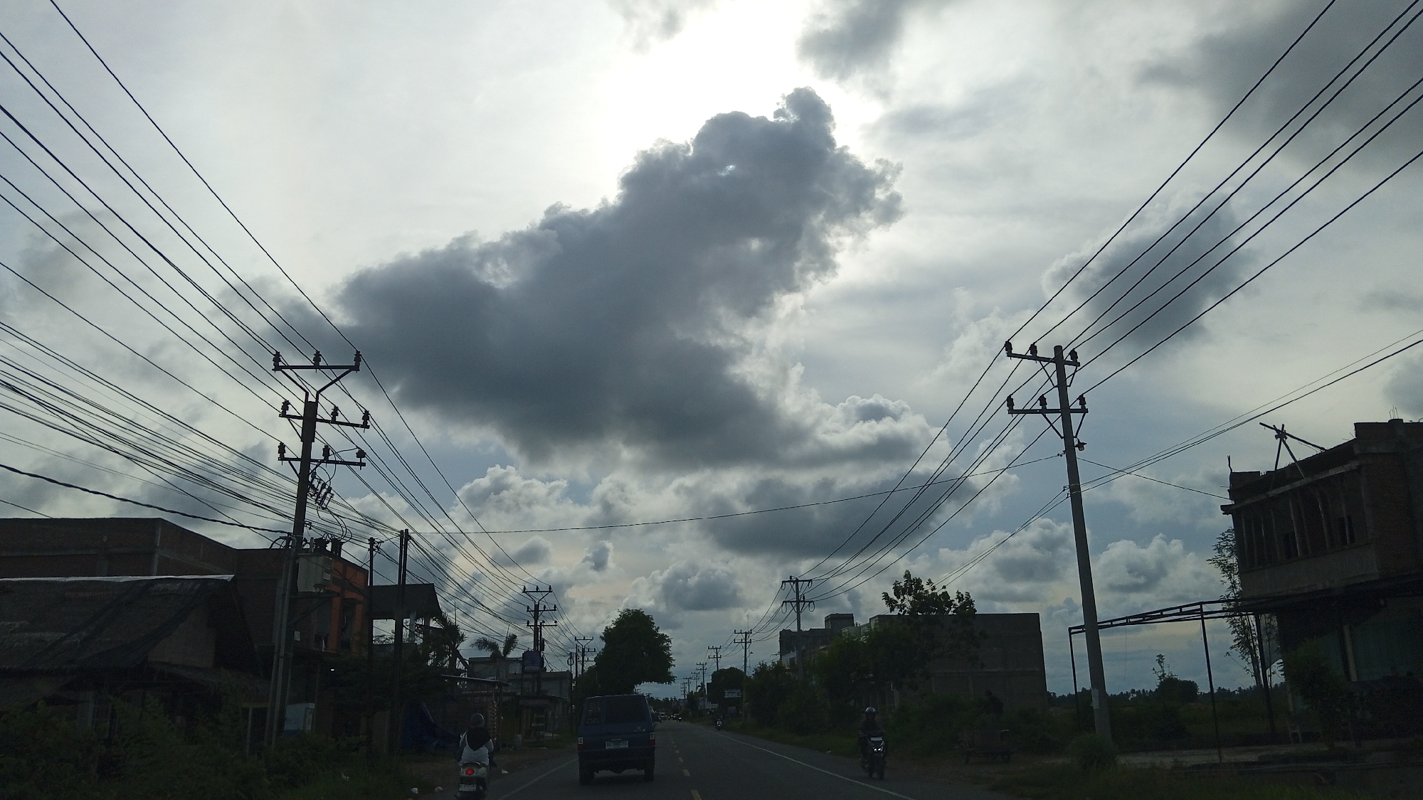 The image depicts a street scene in Payameuneng, Indonesia, under a cloudy sky. A two-lane asphalt road runs through the center of the frame, with a dark blue van driving away from the viewer. Utility poles with numerous wires line both sides of the road. On the left, there are buildings with dark roofs and some construction materials visible. On the right, there are more buildings, some appearing to be under construction. A person on a motorcycle is visible on the right side of the road, also traveling away from the viewer, and another person on a motorcycle is approaching on the left. The overall atmosphere is overcast, suggesting a potentially rainy day.
