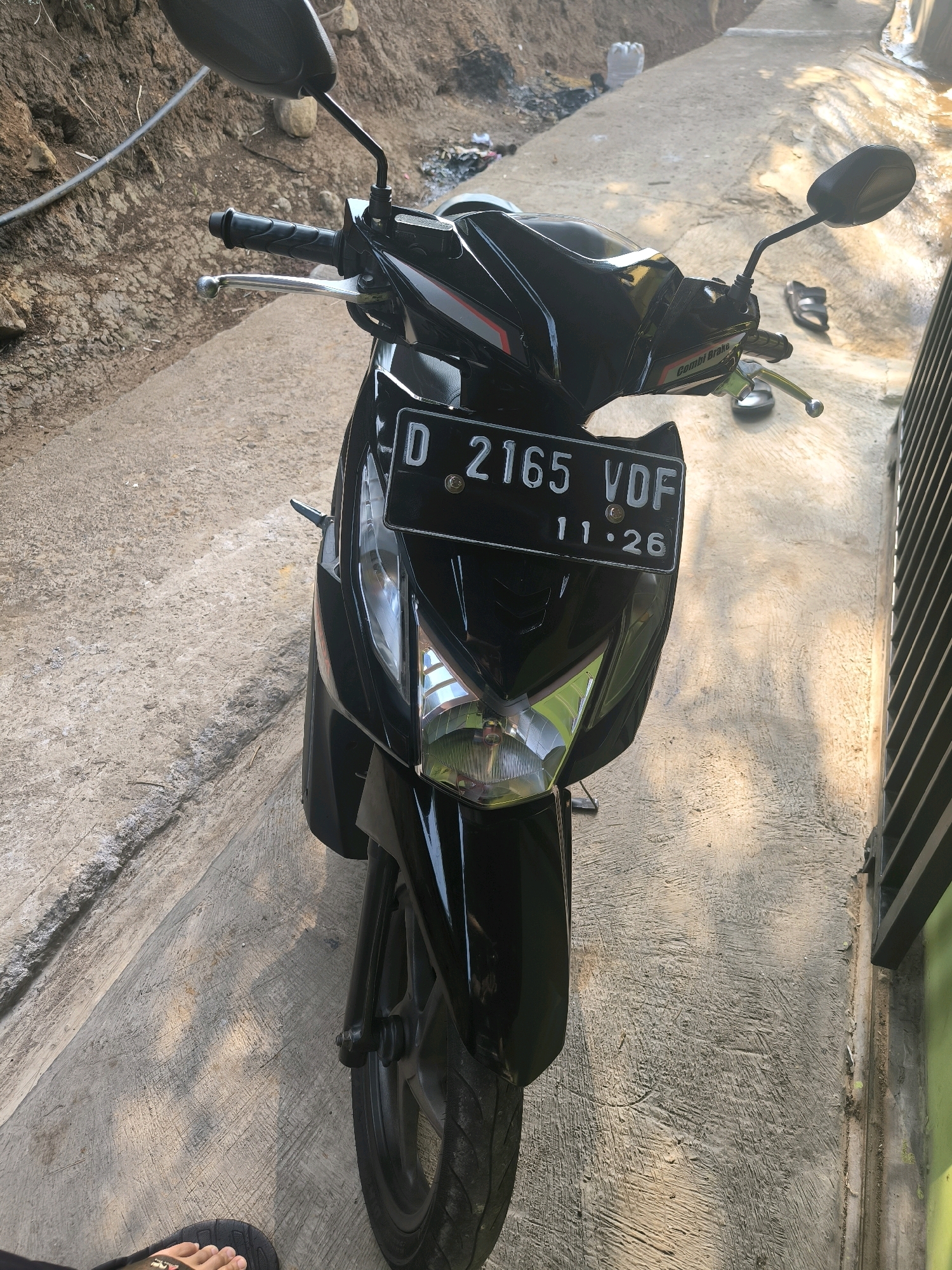 A black motorcycle with a license plate reading "D 2165 VDF 11 26" is parked on a concrete surface outdoors. The motorcycle's front is facing the viewer. The environment appears to be a pathway or road with a dirt embankment on the left and a metal fence or gate on the right. Shadows from the motorcycle and possibly other objects are cast on the ground, indicating sunlight. A person's foot wearing a black sandal is visible in the lower left corner of the image. The location is identified as Cilengkrang, Indonesia.