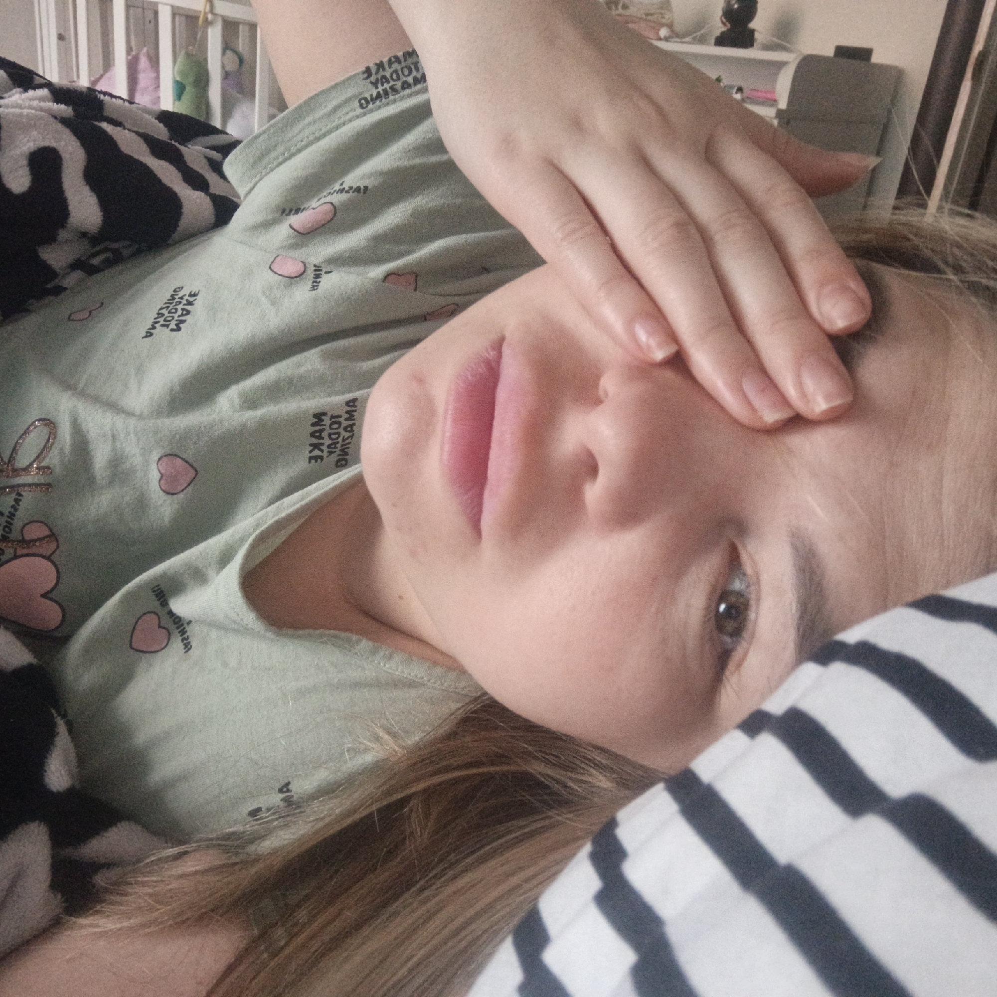 An individual is depicted lying down, possibly in a bed, looking directly into the camera. The person, with light skin and light brown hair, has one hand covering part of their forehead and eyes. They have full, pink lips and one brown eye is visible.
The person is wearing a light green t-shirt or pajama top adorned with small pink hearts and repeated, mirrored text. The visible text, when un-mirrored, reads "MAKE TODAY AMAZING." A black and white zebra print blanket covers part of their body, and a black and white striped pillow or blanket is visible in the foreground near their head.
In the background, a white, slatted structure resembling a crib or headboard is present, with what appears to be soft toys or clothing hanging from it. Further back, a shelf with a dark object and other indistinct items can be seen. The lighting is soft and ambient, suggesting an indoor setting. The image is taken from an angled perspective, with the person lying on their side relative to the camera's view.