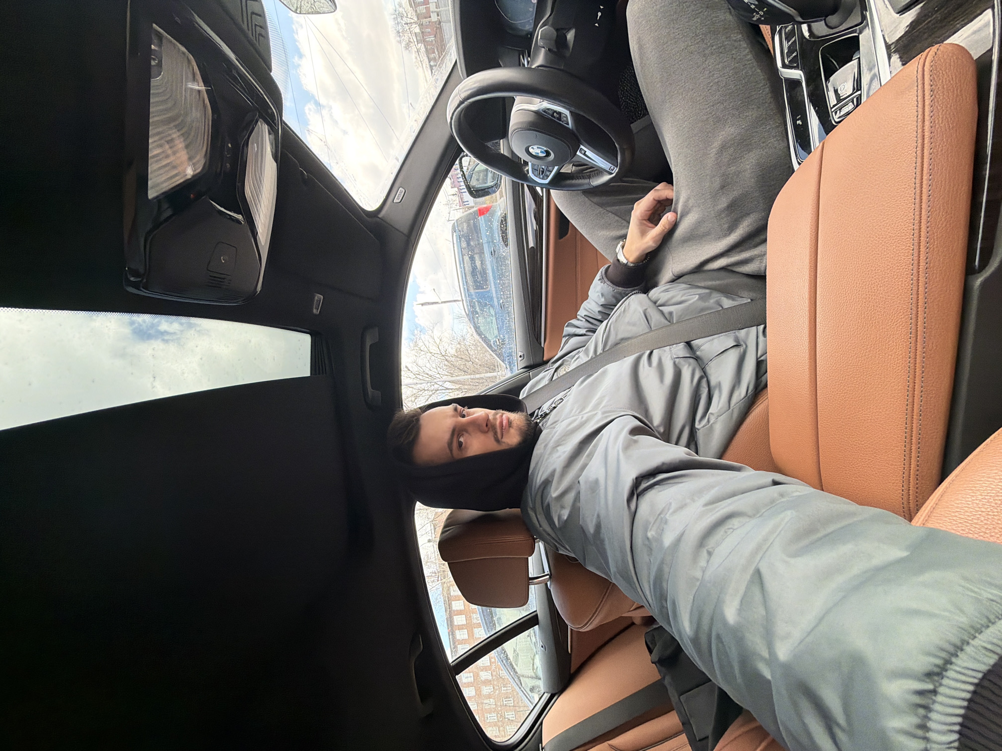 A man is sitting in the driver's seat of a BMW car. He is wearing a gray jacket and a dark hood. The car has brown leather seats and a sunroof. Outside the car, buildings can be seen, indicating an urban environment. The location is Krasnoyarsk, Russia.