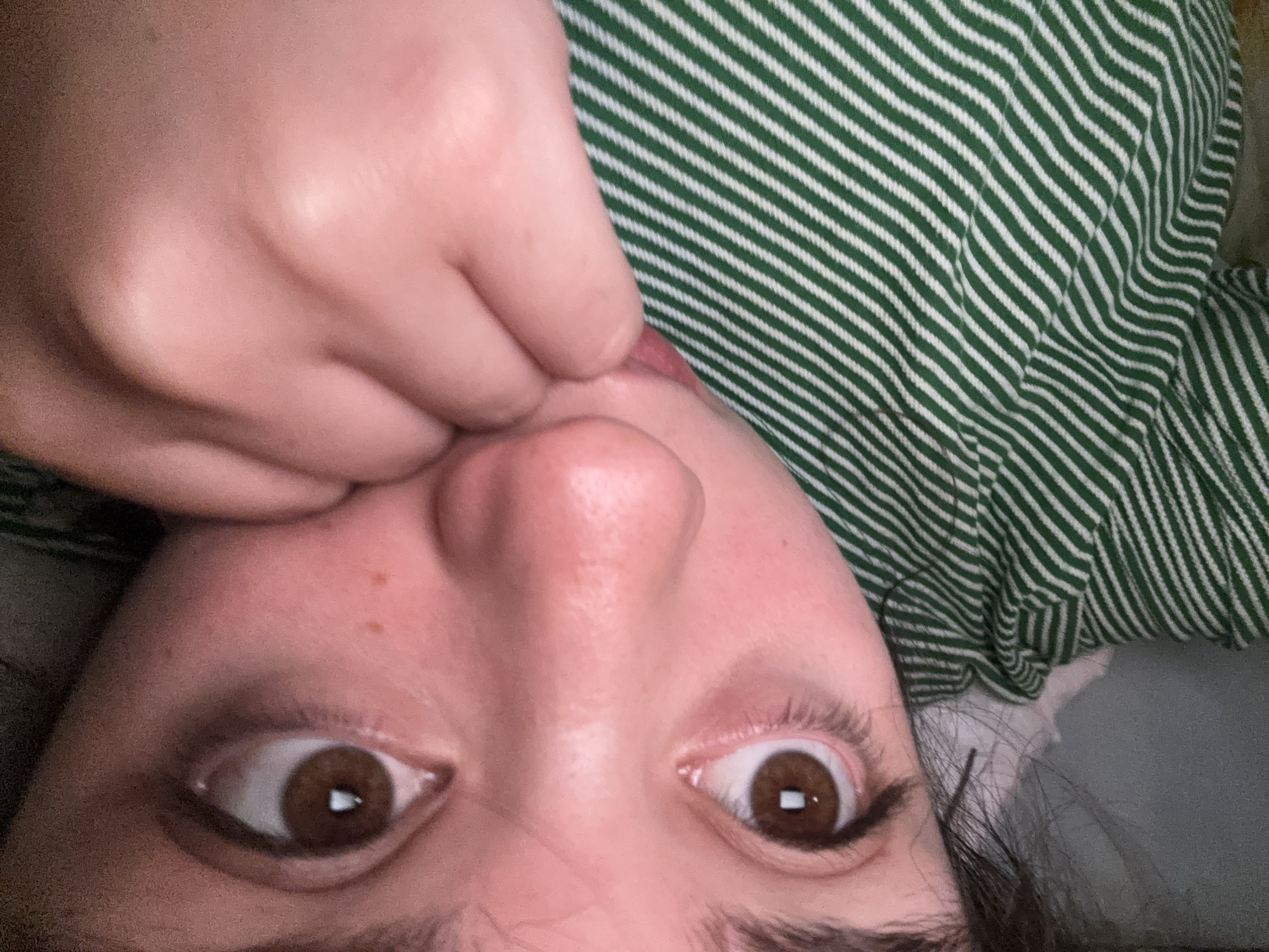 The image shows a close-up, inverted perspective of a person's face, with their fingers near their mouth. The person has brown eyes and light skin. They are wearing a green and white striped shirt. The environment is indeterminate. The image does not contain enough information to establish a connection to Garmen, Bulgaria.