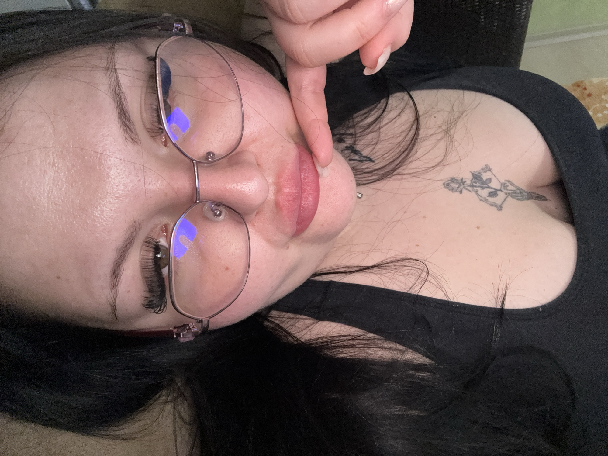 The image shows a woman with glasses and a tattoo on her chest. Her finger is touching her lips. The location is not discernible from the image.