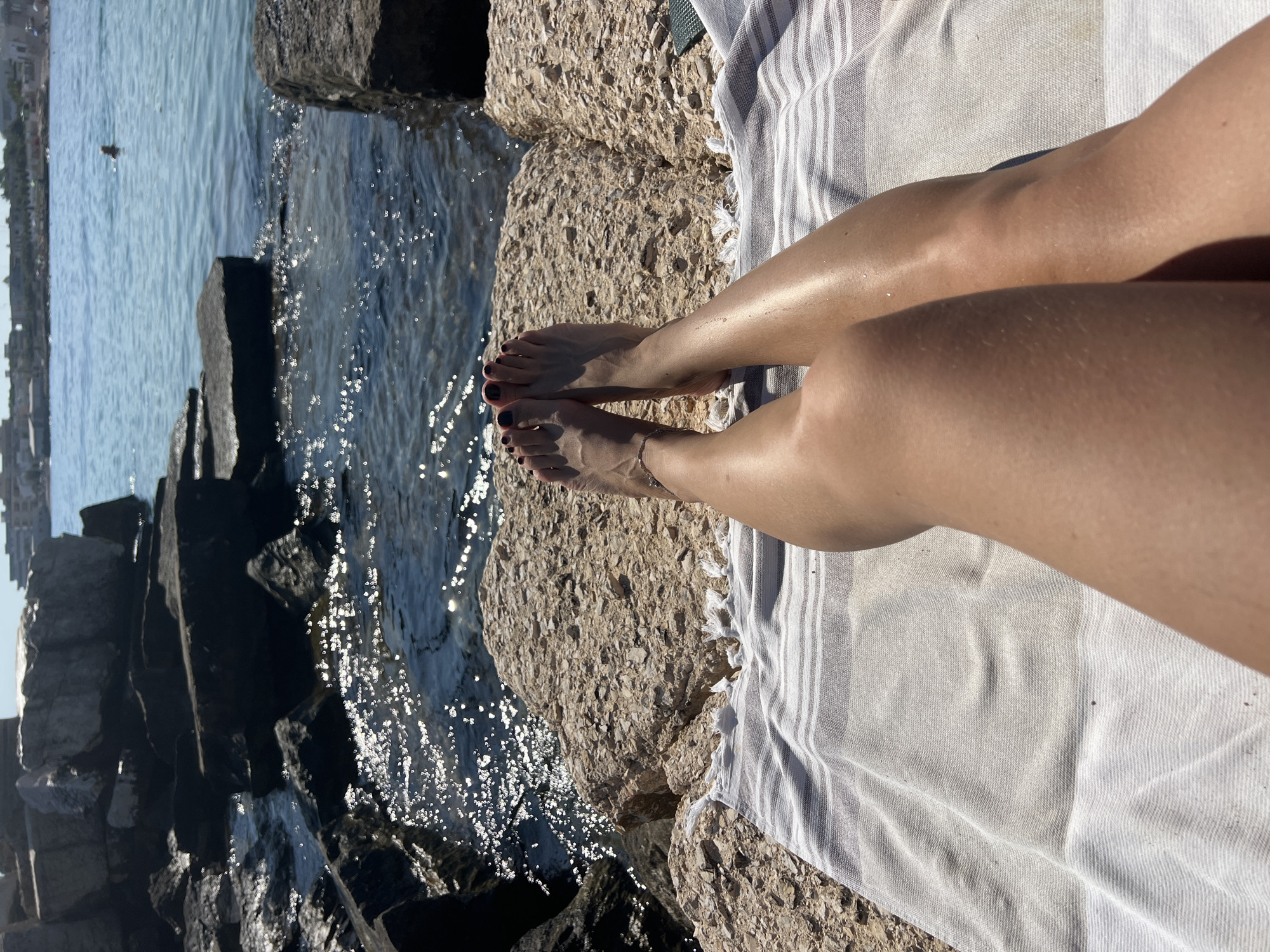 A first-person perspective shot shows a person's legs and feet relaxing on a striped towel placed on a rocky shore, with the sea in the background. The sun is shining, casting shadows and creating a sparkling effect on the water. In the distance, the coastline of Bari, Italy, is visible, with buildings and a few people near the water. The overall scene conveys a sense of leisure and vacation.