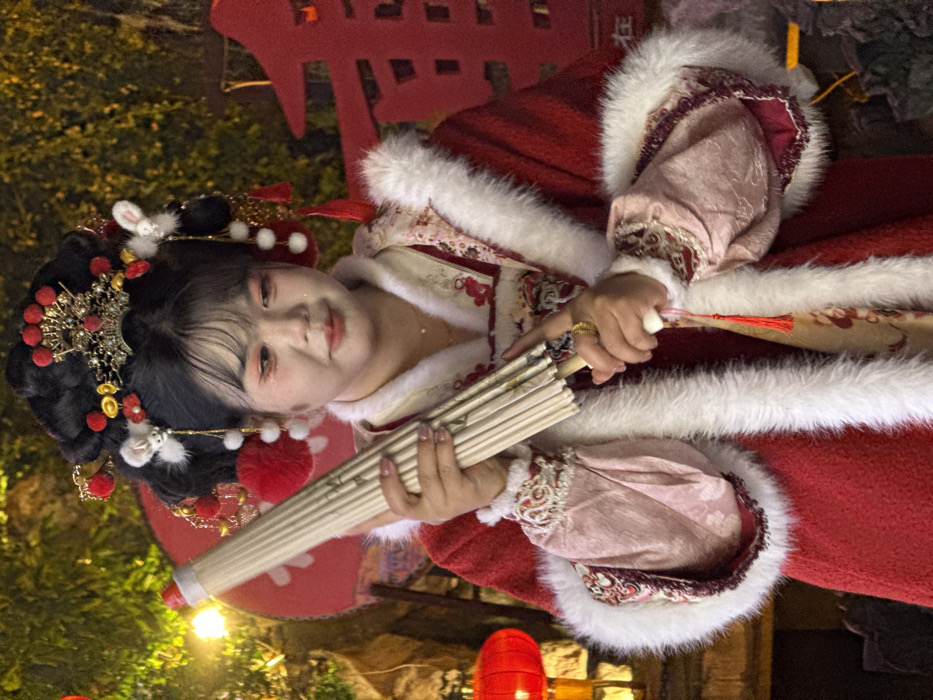 A medium-shot image depicts a person, appearing female, standing in an outdoor or semi-outdoor festive setting in Yuzhong District, China. The individual, positioned slightly off-center to the left, is wearing elaborate traditional Chinese attire, primarily a thick red overcoat with extensive white fur trim along the collar, front opening, and cuffs. An inner garment with intricate patterns, possibly in shades of pink and beige, is visible beneath the overcoat, particularly on the sleeves.
The subject has dark hair styled into an ornate updo adorned with a gold-colored headdress featuring red gems, multiple red pompoms, white fluffy rabbit-shaped ornaments, and gold dangling chains. Makeup includes prominent reddish-pink eyeshadow on the eyelids and under the eyes, and light lip color. The individual holds a closed, light-colored, possibly off-white or beige, paper or silk parasol with a wooden handle in both hands, angled upwards and to the left. A small red tassel hangs from the parasol's handle. A gold-colored ring is visible on the ring finger of the right hand.
The background is dimly lit and blurred, suggesting an evening or night scene. To the left, a bright red lantern is visible, partially obscured by illuminated green foliage. To the right, a large red decoration with partially visible Chinese characters is present. The overall environment suggests a cultural or celebratory atmosphere.