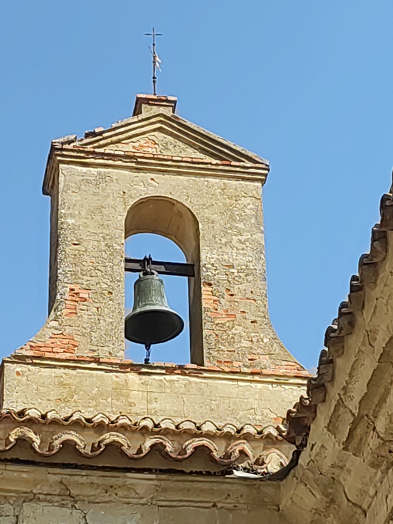 The image captures a close-up, low-angle view of a bell tower and adjoining roof structure against a clear blue sky. The bell tower is constructed of weathered stone with exposed brickwork, topped with a triangular pediment and a small cross. A dark metal bell is suspended within an arched opening in the tower. The roof features terracotta tiles arranged in a wavy pattern. To the right, another section of a stone building with a textured, decorative roof edge is visible. This scene is located in Ampudia, Spain, specifically depicting the historic bell tower of the Church of San Miguel Arcángel. There are no people or animals visible in the frame, nor any observable actions or interactions. The environment is bright and sunny, with no indication of adverse weather.