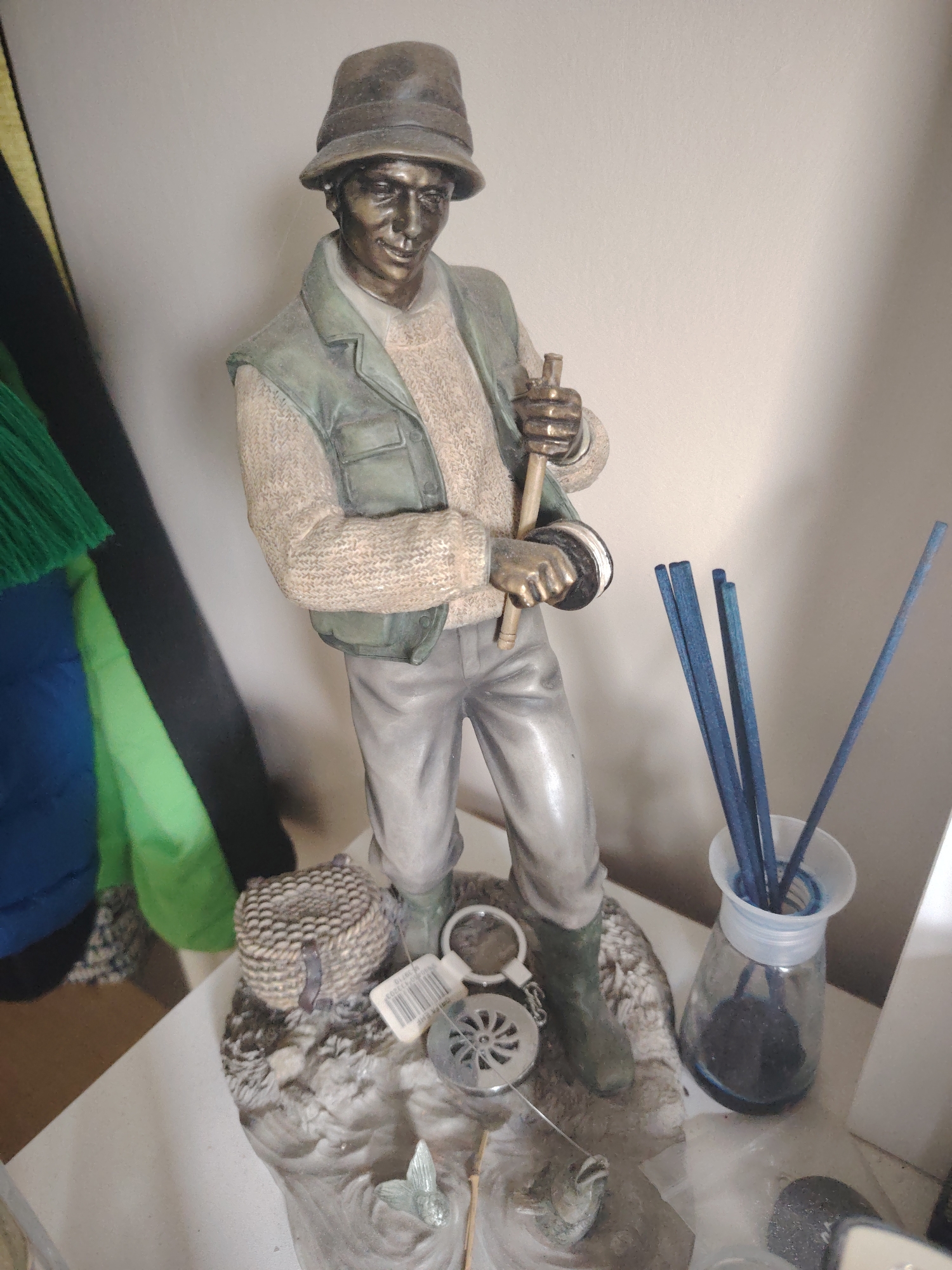 A bronze-colored statue of a male fly-fisherman stands on a light-colored surface in Huntingdonshire, United Kingdom. The statue depicts a man wearing a bucket hat, a textured light-colored sweater, a green fishing vest, grey trousers, and dark green boots. He holds a fishing rod with a wooden shaft and a fly reel. The statue's base is textured to resemble a natural landscape with rocks and water, featuring a small woven fishing creel, a silver-colored landing net, a compass-like object with a white barcode tag, and a fish with a fishing line extending from its mouth. To the right of the statue, a glass diffuser bottle containing dark liquid holds several blue reed diffuser sticks. The background consists of a plain, light-colored wall, with multiple colored fabrics (yellow, black, green, blue) partially visible on the left side, casting a shadow on the wall.