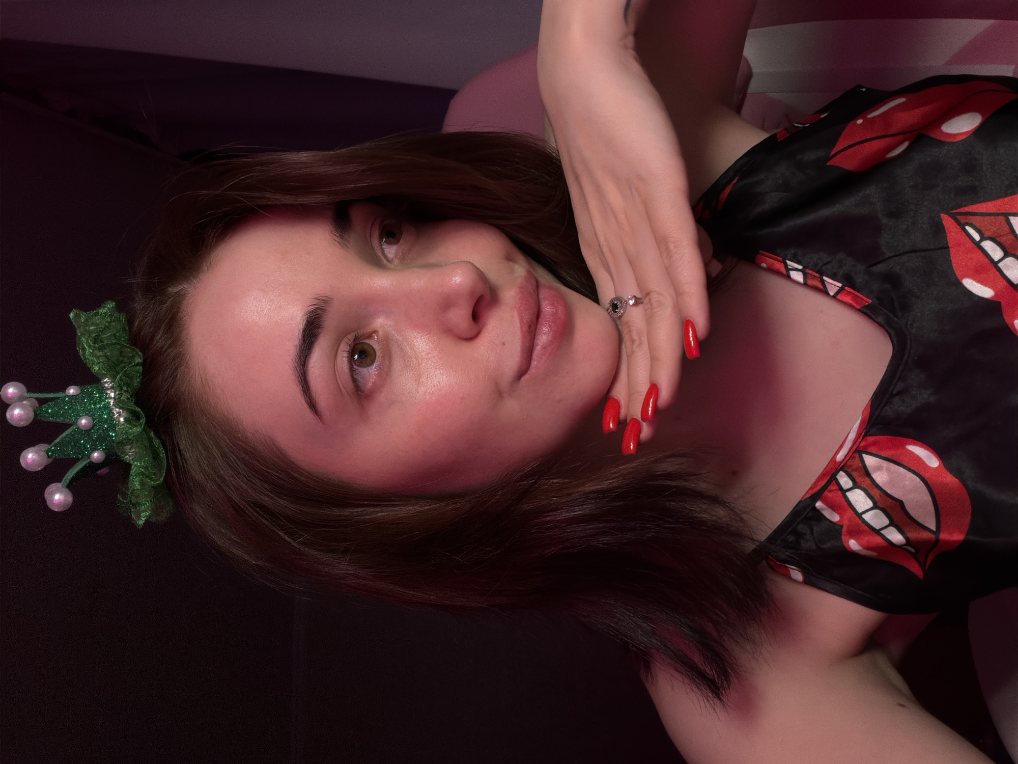 A young woman with brown hair and brown eyes is captured in a close-up, head-and-shoulders portrait. She is wearing a black camisole with a bold red and white lip print pattern. A sparkly green crown with white pearl-like accents rests on top of her head. Her right hand is gently supporting her chin, and her fingernails are painted a vibrant red. She wears a silver ring with a dark stone on her right ring finger. The background is dark and softly lit, with hints of pinkish-purple light casting a warm glow on her skin. The overall impression is one of playful self-portraiture.