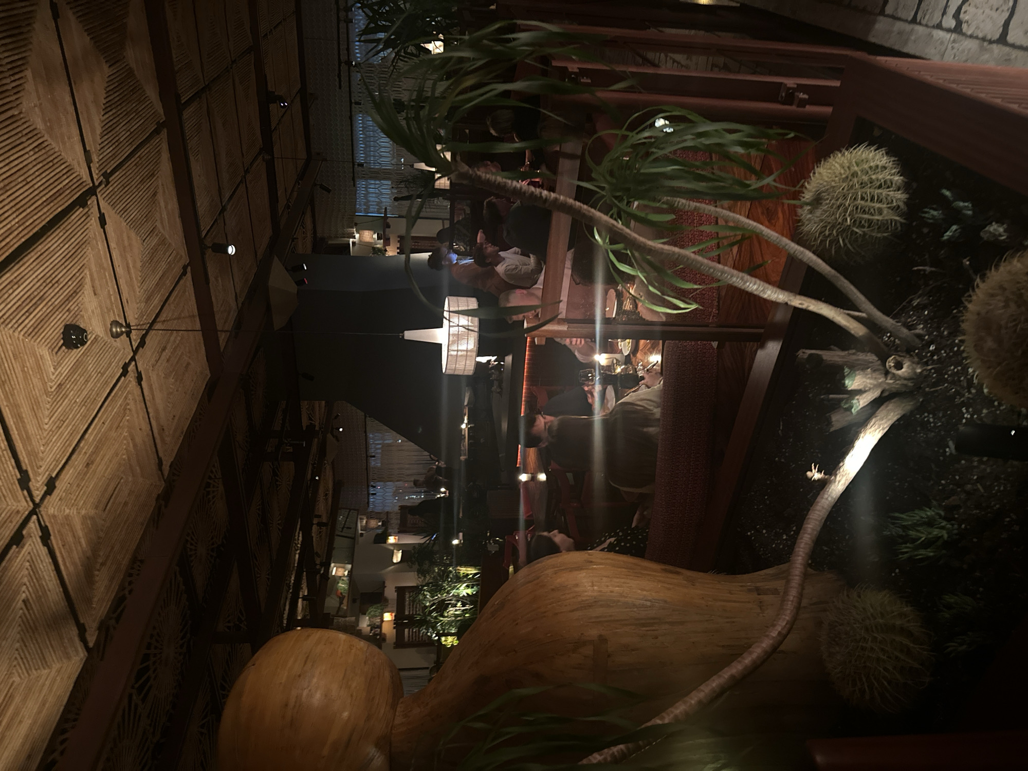 The image captures the interior of a dimly lit restaurant. Patrons are seated at tables enjoying meals. In the foreground, a large, possibly wooden, sculpture stands prominently, accompanied by cacti and various plants in containers. The restaurant features distinctive woven ceiling patterns. The exact location within London, United Kingdom, cannot be determined from this image alone.