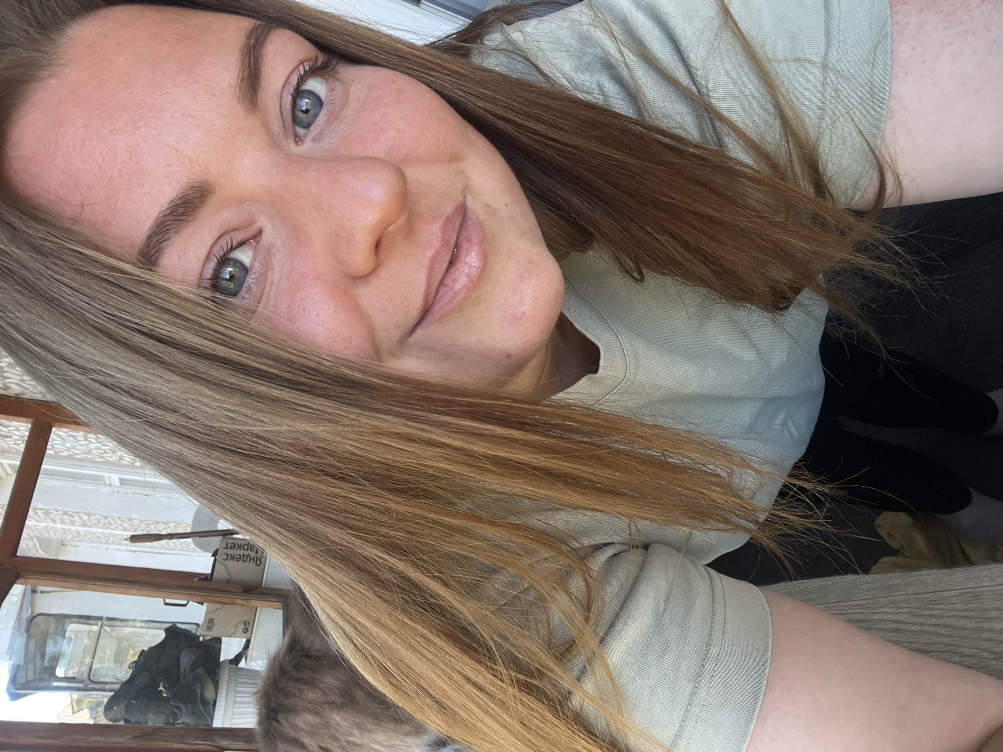 A woman with light brown hair and light complexion is posing for a selfie. She is wearing a light-colored t-shirt. The background shows a window with a view that includes a white truck, boxes, and miscellaneous items. There is no definitive evidence within the image to confirm the location in Razdolie, Russia.