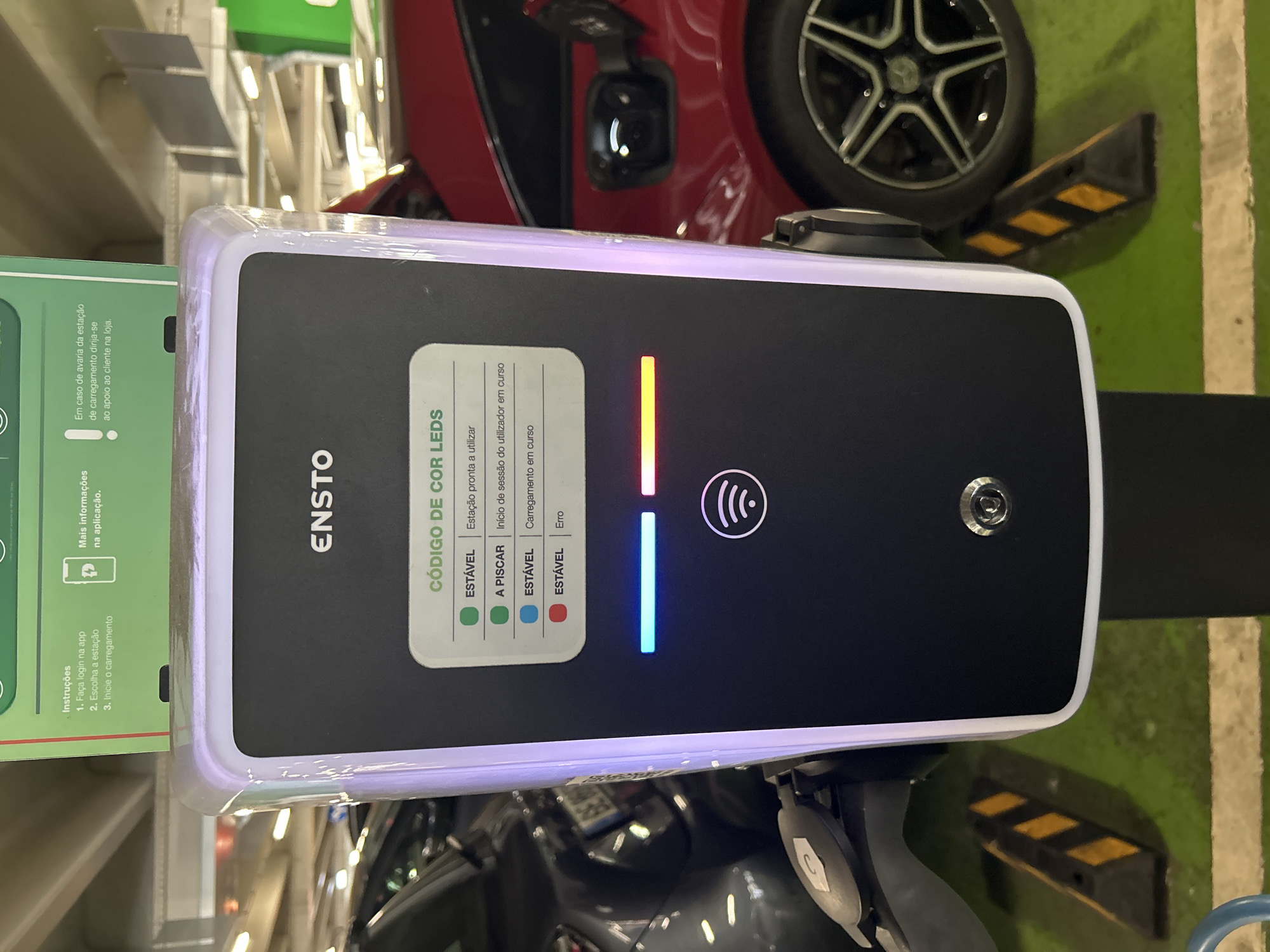 A full-shot, eye-level, outdoor, eye-level, vertical photograph captures an Ensto electric vehicle charging station in a parking garage. The charging station is positioned centrally and illuminated with white and purple light around its edges. It features a black screen displaying information in Portuguese about LED indicators, with options for "Estável" (Stable) and "A piscar" (Blinking). Below the screen are illuminated LED bars: a blue one on the left, a yellow one in the middle, and a red one on the right. A Wi-Fi symbol is displayed beneath the LED bars.
To the left of the charging station, a portion of a dark-colored car is visible, and to the right, a red car is parked. The ground is covered in green asphalt with yellow and black striped parking guides. The scene appears to be in the parking area of a shopping center, potentially in Senhora da Hora, Portugal, as indicated by the presence of the Ensto charging station, a brand commonly found in Europe. There are no people present in the frame. The overall lighting suggests it is daytime, with artificial lights from the parking garage also contributing.