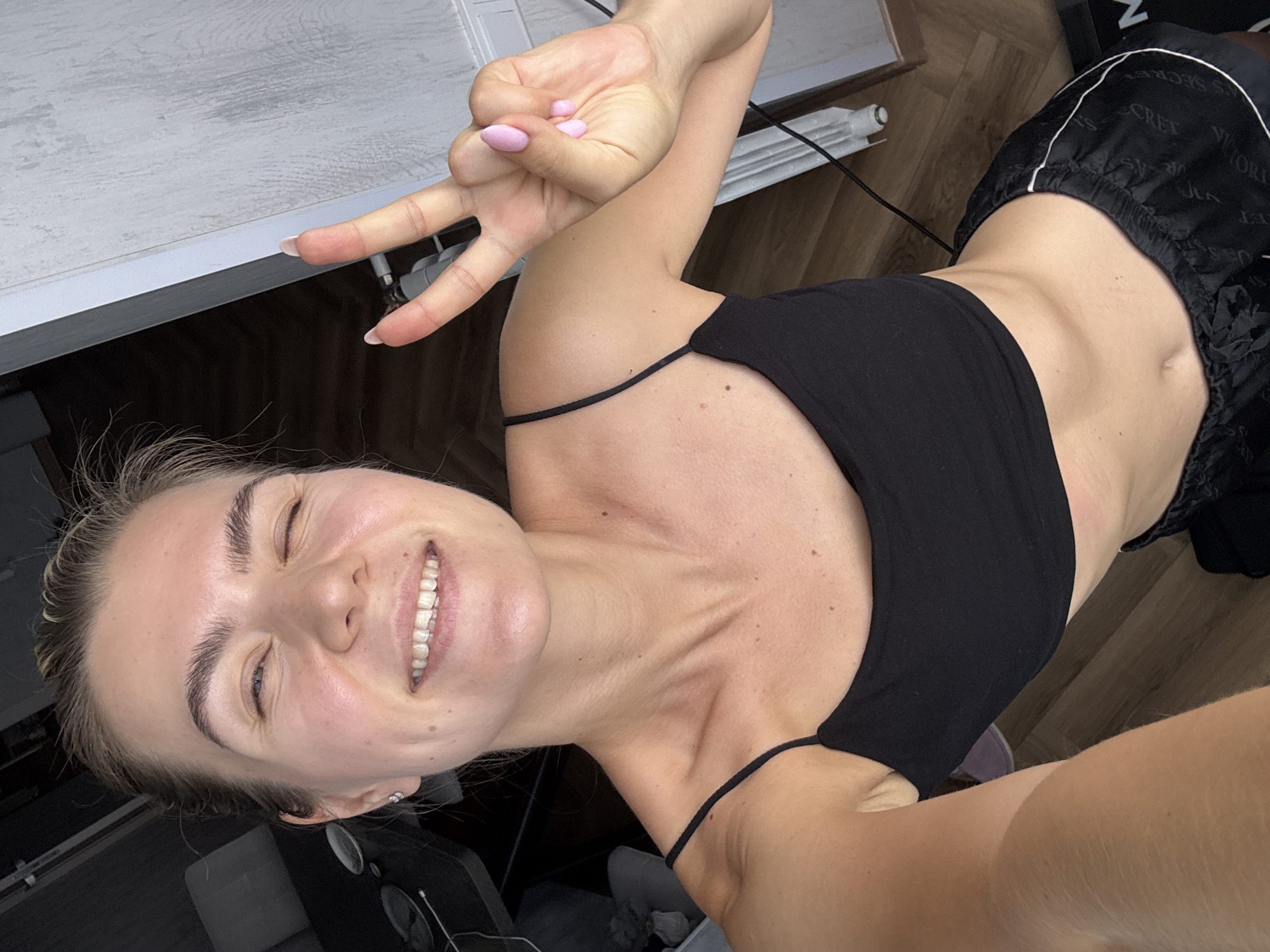 This media file is a close-up, high-angle selfie of a young woman. She is smiling and winking, with her right hand held up in a peace sign. Her hair is blonde and pulled back. She is wearing a black tank top and black shorts with white lettering that appears to be "VICTORIA SECRET". The setting appears to be indoors, possibly a bedroom or living area, with a wooden floor visible. The lighting suggests it could be daytime. The overall mood is cheerful and playful.
