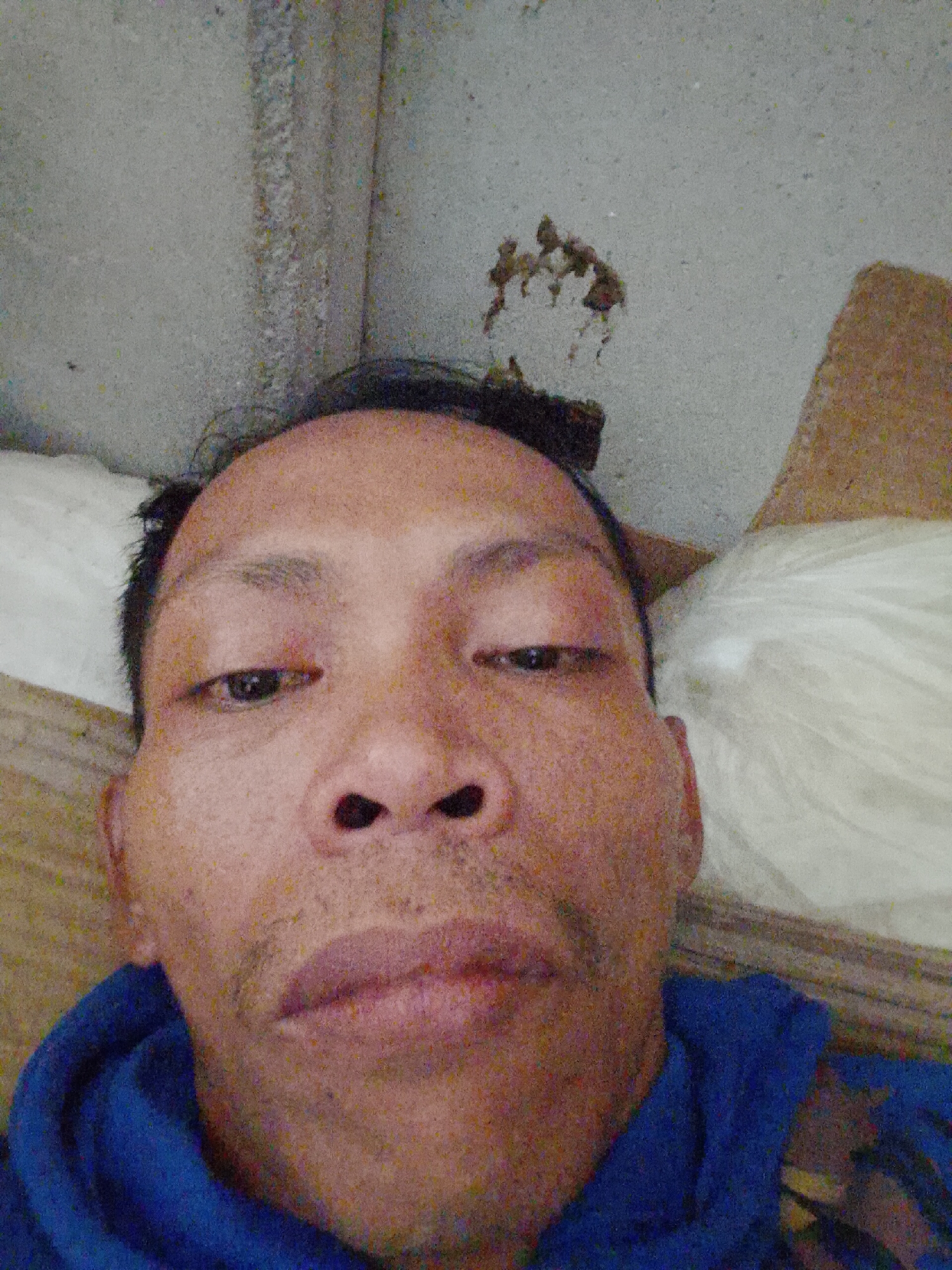 A close-up selfie of an Asian male with short, dark hair and a blue hooded sweatshirt. He appears to be lying down, possibly on a wooden surface with a white cushion behind him. The background shows a concrete wall with some discoloration. It is impossible to verify the location of Bukit Ketok, Indonesia from the image.