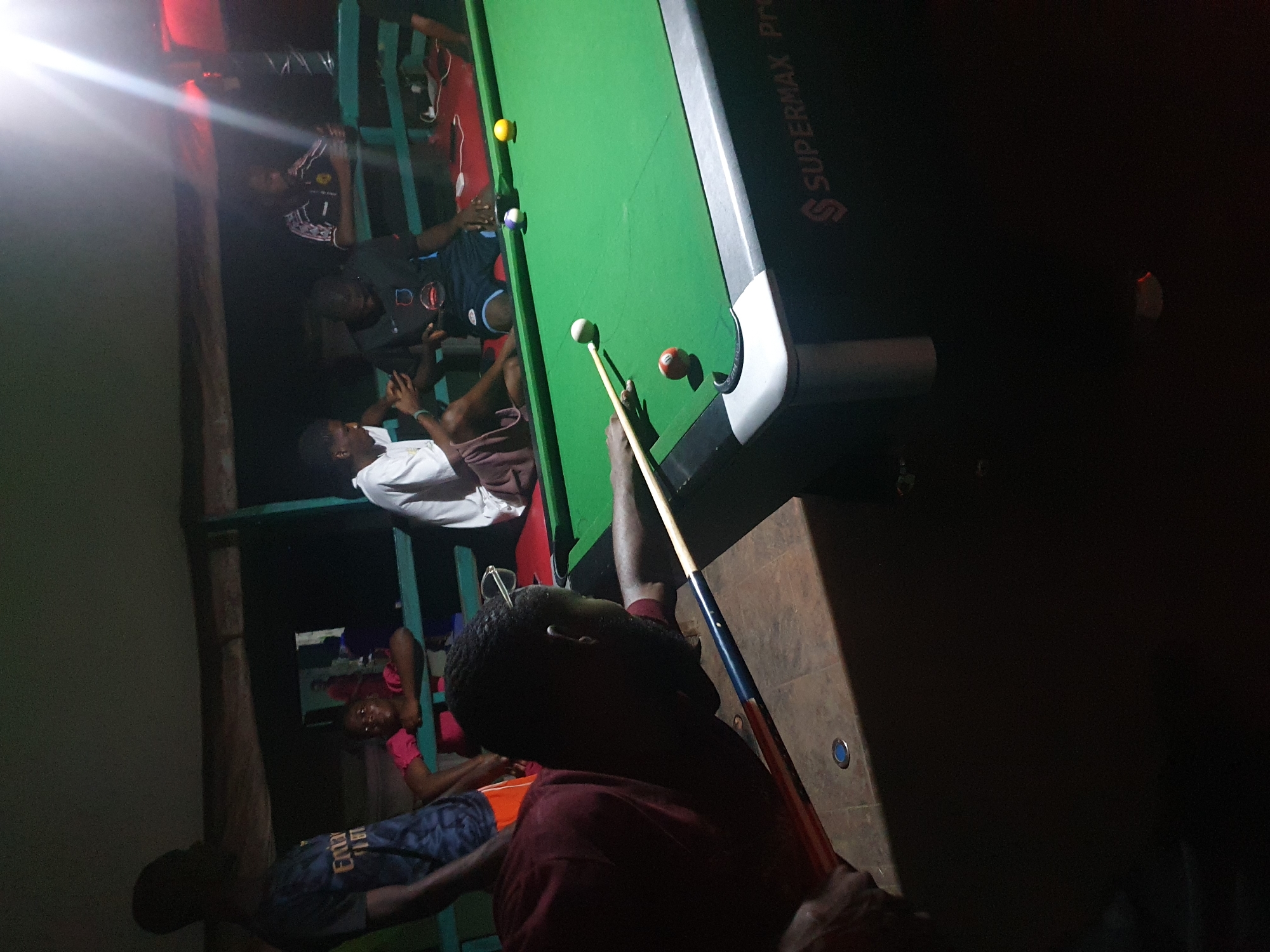 A group of young men and a woman are gathered around a pool table in what appears to be a dimly lit outdoor or semi-outdoor setting. The pool table is green with several balls on it, including a white cue ball, a striped ball, and a solid yellow ball. One of the men, in the foreground and facing away from the camera, is in the process of striking the cue ball with a pool cue. He is wearing a dark red t-shirt. Other individuals are seated or standing around the table, observing the game. One woman in a pink top is leaning on a green railing. Another man is wearing a dark t-shirt with a pattern and shorts. The environment suggests a casual gathering, possibly at a recreation center or a private residence in Lokoja, Nigeria, given the available contextual information. The lighting is dim, with a strong light source visible in the upper right corner of the image, casting bright reflections. The overall atmosphere seems relaxed and social.