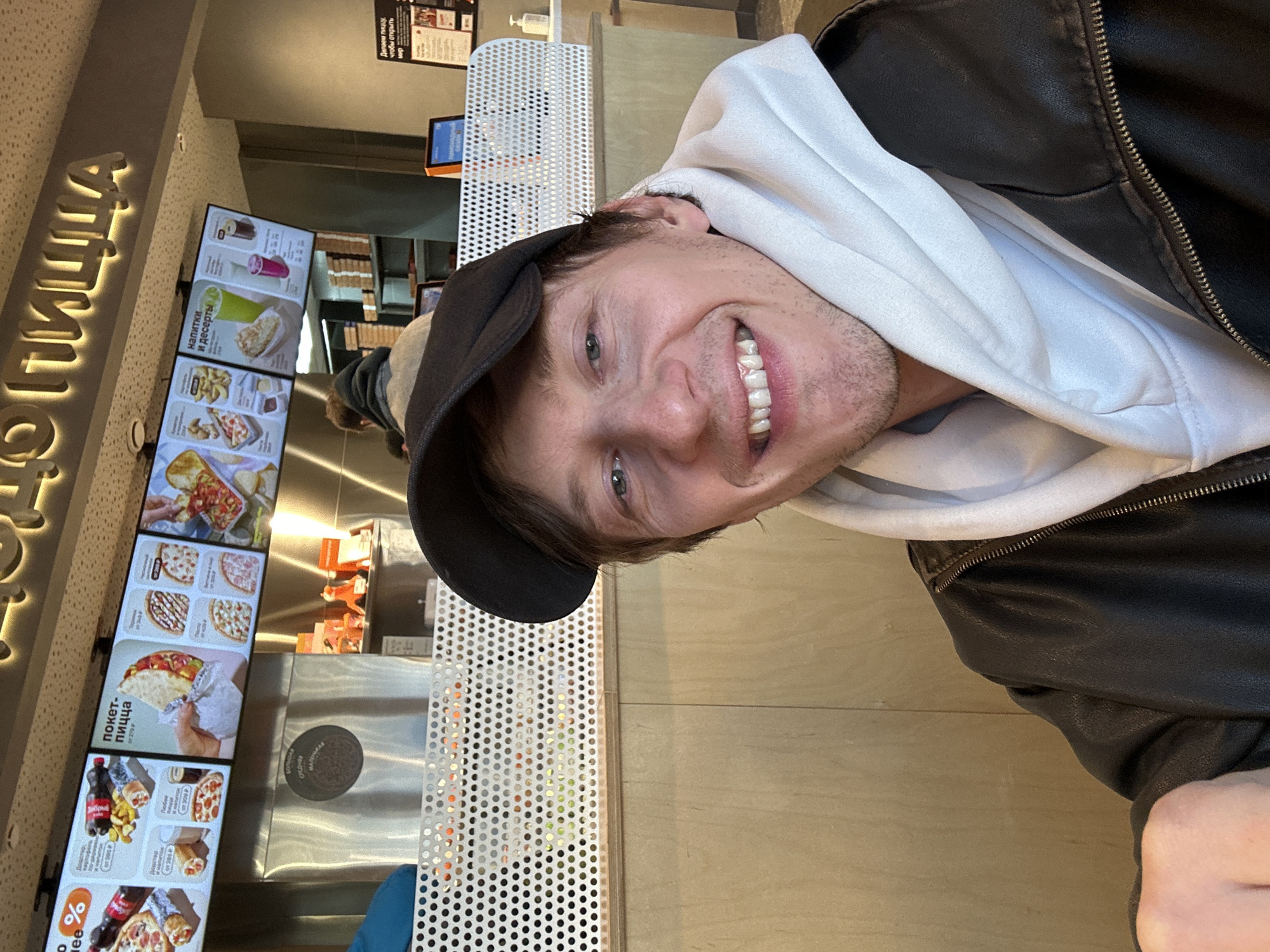 A young man with fair skin, light-colored eyes, and light stubble smiles broadly at the camera in what appears to be an indoor fast-food restaurant or pizzeria in Murino, Russia. He is wearing a black baseball cap, a white hoodie, and a black leather jacket. His expression is cheerful and happy.
The setting is a brightly lit, modern establishment with light-colored walls and wooden-textured surfaces. In the background, to the left, multiple digital menu screens display various food and drink items, including different types of pizza, "покет-пицца" (pocket-pizza), "напитки и десерты" (drinks and desserts), and prices. A large, illuminated Cyrillic sign on the wall spells out "ПИЦЦА" (PIZZA), part of what is likely a brand name such as "Додо Пицца," which is also visible on one of the menu screens. Another slogan seen on a small sign reads: "Делаем пиццу, чтобы открыть мир" (We make pizza to open the world).
A counter area with a distinctive white perforated metal panel is visible. The man is positioned with this counter to his side. The overall atmosphere suggests a casual dining experience, likely during the daytime given the bright interior lighting.