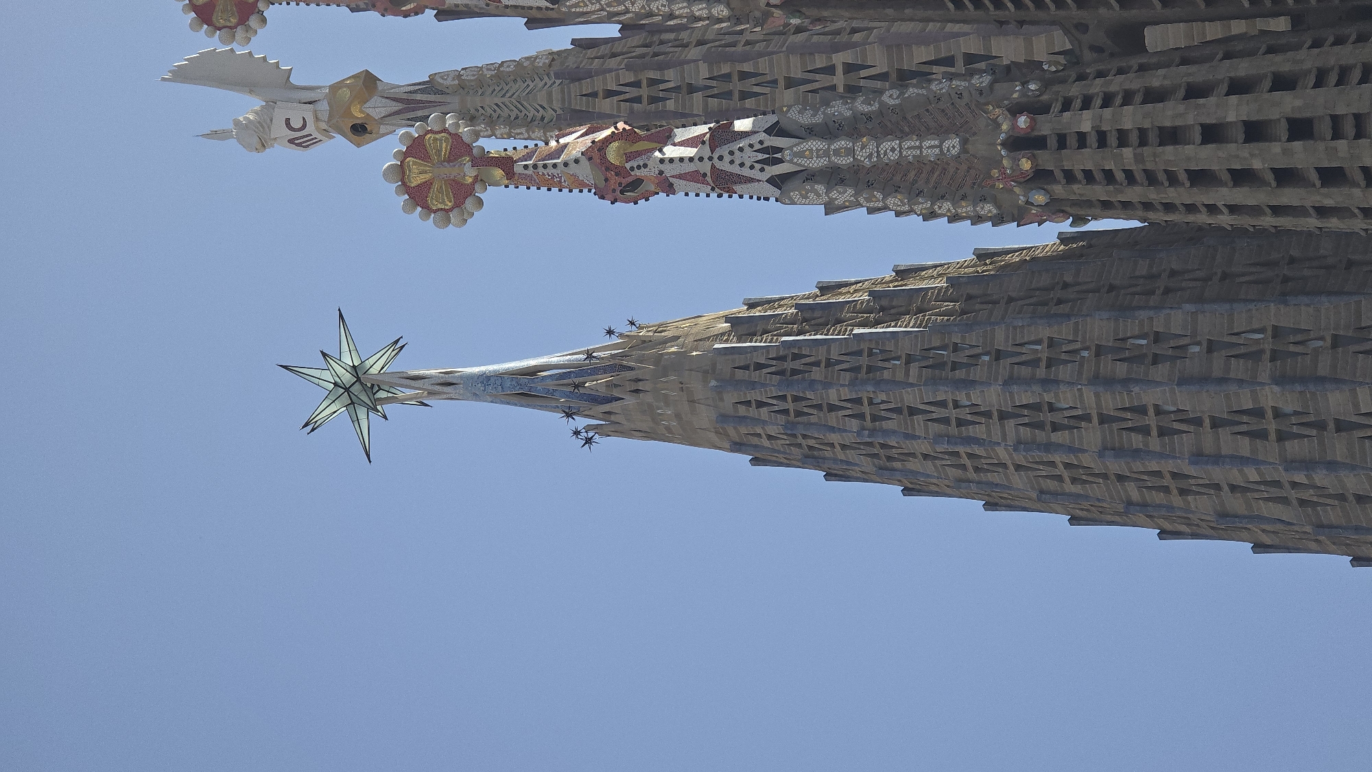 The image shows a close-up, low-angle view of the spires of the Sagrada Familia basilica in Barcelona, Spain. The sky is a clear, bright blue. The central spire in the foreground is tall and conical, made of stone with a repeating pattern of triangular openings. It is topped by a large, translucent, star-shaped ornament. To the right, another spire, adorned with intricate, colorful mosaic work and sculptural details, rises higher. A third spire, partially visible on the far right, also displays similar decorative elements. The architecture is characteristic of Antoni Gaudí's unique style, known for its organic forms and religious symbolism. There are no people visible in the frame.