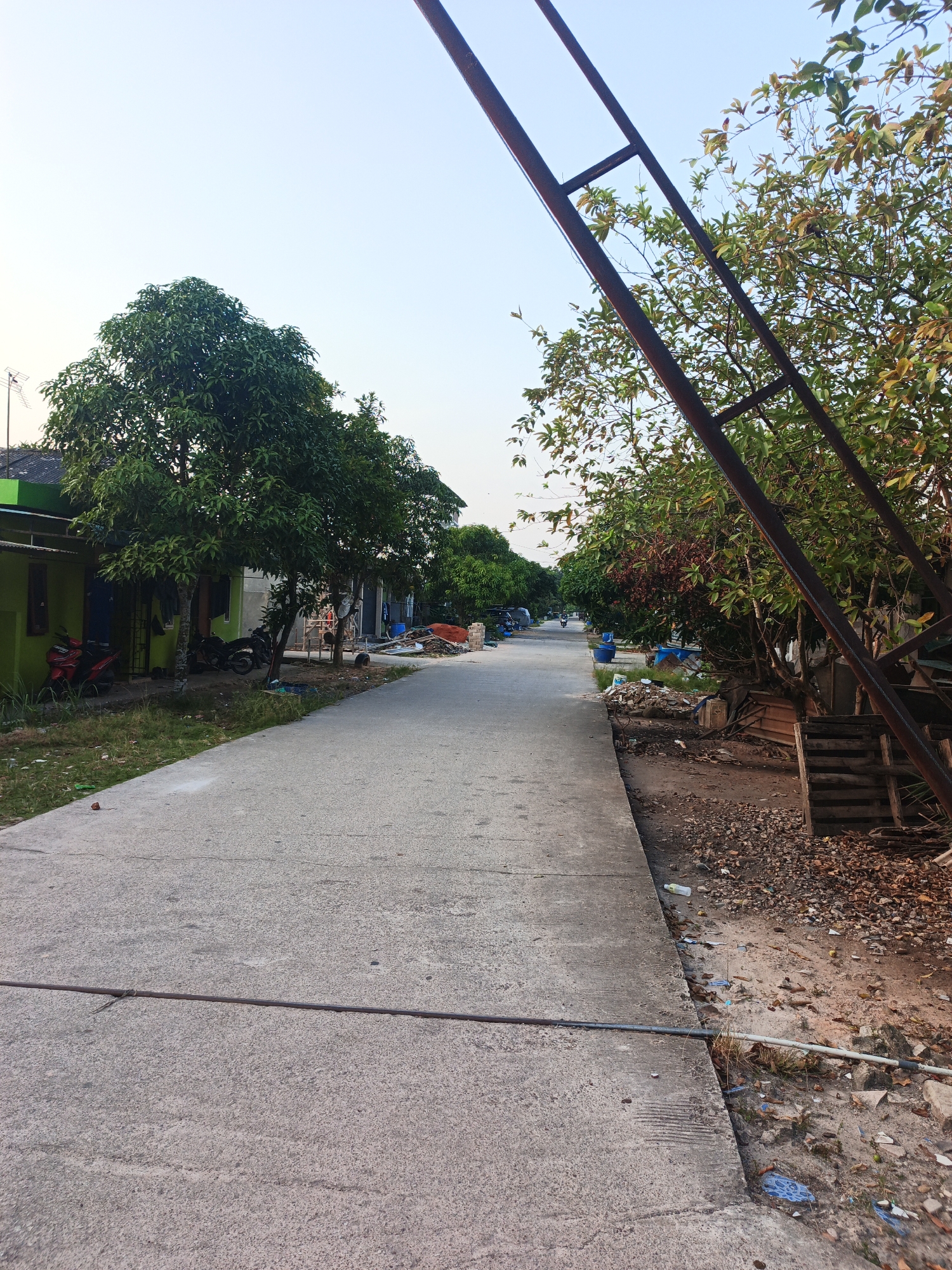 The image depicts a street scene in a residential area, possibly within the city of Batam, Indonesia. The perspective is from ground level, looking down a paved road lined with modest houses on the left and vegetation and some construction materials on the right. The sky is clear and light blue, suggesting it is either early morning or late afternoon.
On the left side of the road, several houses with green and gray exteriors are visible. Some trees with dense green foliage are planted in front of these houses. Parked on the side of the road are two motorcycles, one red and one black. Clothes are hanging on a line in front of one of the houses.
The right side of the road features a mix of trees, some with reddish-brown leaves, and a visible wooden structure, possibly part of a construction or storage area. There are piles of debris, including leaves, wood scraps, and plastic items, scattered along the edge of the road. Two large blue barrels are also present.
In the distance, the road continues and appears to curve slightly to the left. More houses and trees are visible further down the street, creating a sense of a residential neighborhood. A single motorcycle is seen riding in the distance on the road. A prominent dark metal structure, likely part of a large gate or frame, extends diagonally across the upper right portion of the image. The ground in the foreground is paved concrete, with a darker metal rod running horizontally across it, secured with a tie.
