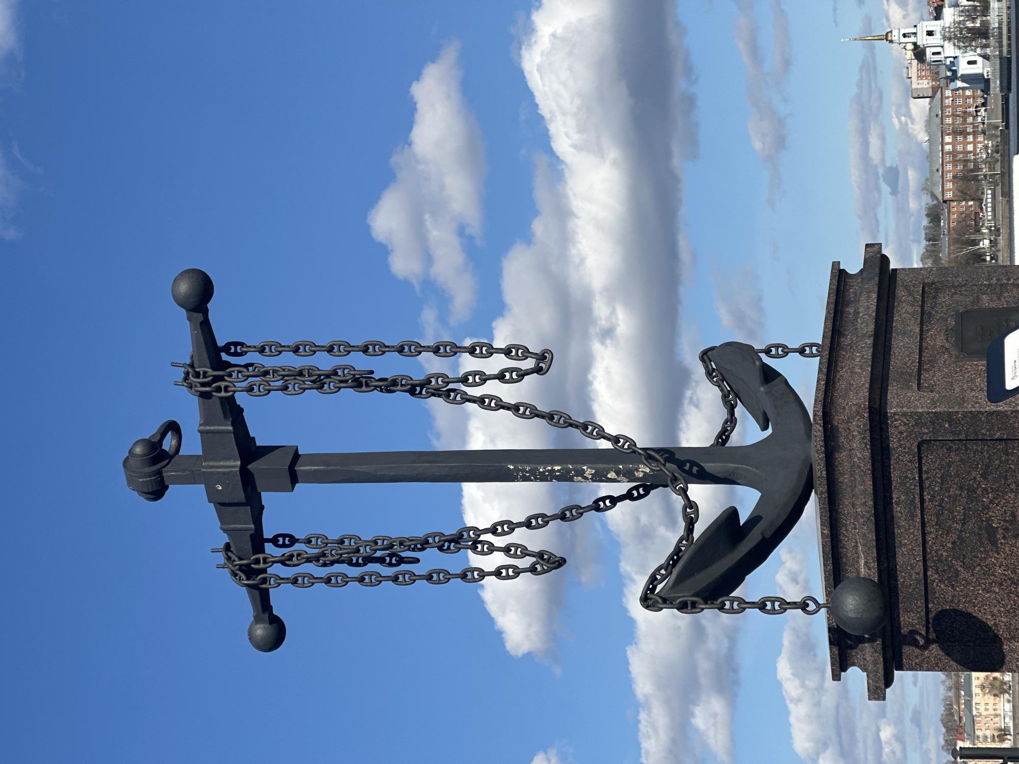 The image shows a monument in Votkinsk, Russia. The monument consists of a dark anchor draped with heavy chains. The anchor is mounted on a tall granite pedestal with spherical ornaments. Behind the monument, the sky is blue with white clouds. In the background, buildings can be seen in the distance.