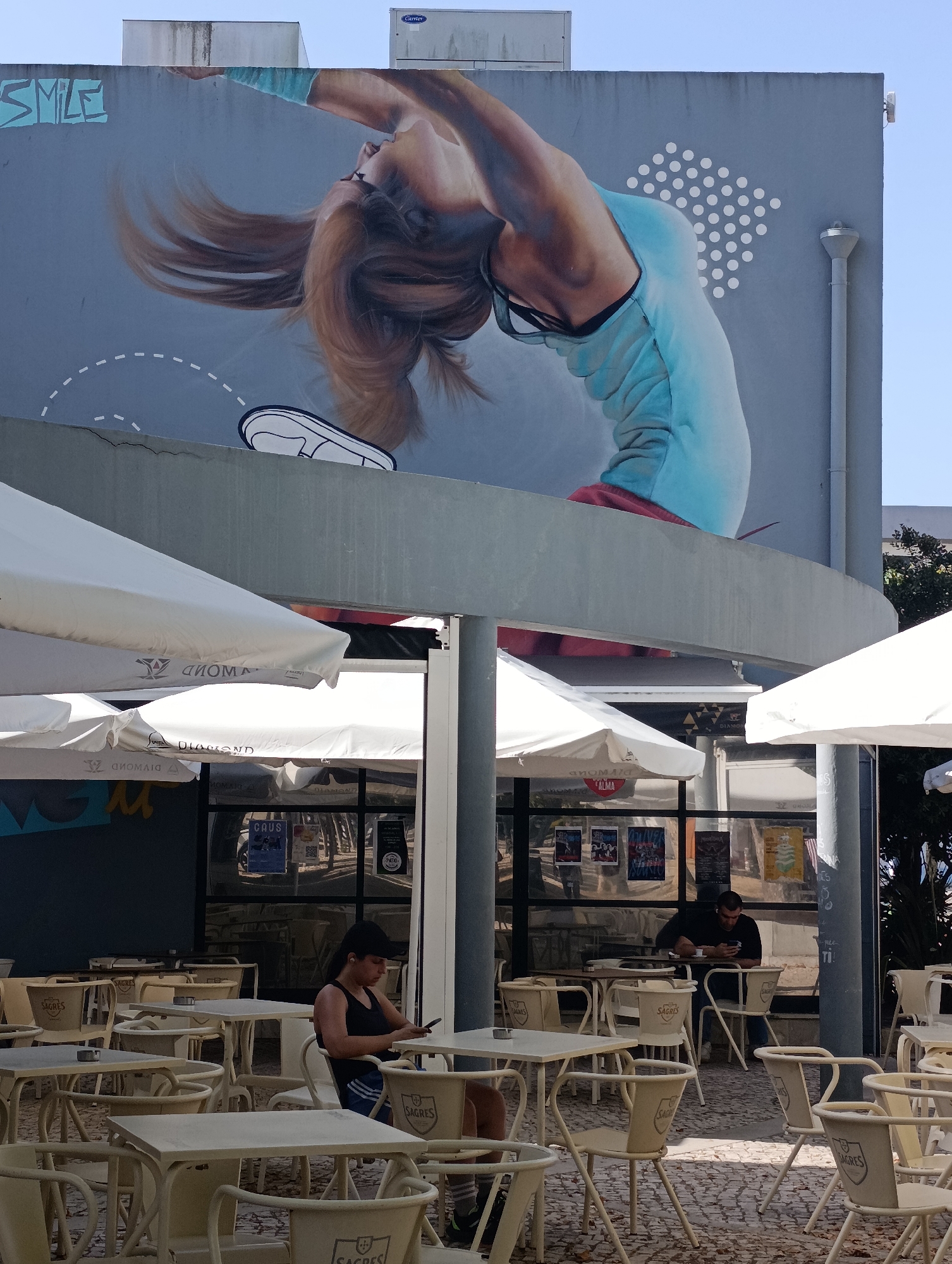 A wide shot of an outdoor cafe scene in Setúbal, Portugal, shows a large mural of a woman performing a backbend on a building facade. The mural is bright and colorful, with the woman's hair flowing and her body in a dynamic pose. Below the mural, several white umbrellas cover tables and chairs arranged on a cobblestone patio. Two men are seated at tables, one in the foreground looking at his phone and another in the background also looking at his phone. The cafe appears to be a popular spot for outdoor dining, with many tables and chairs set up. The image is taken on a sunny day, with clear blue skies.