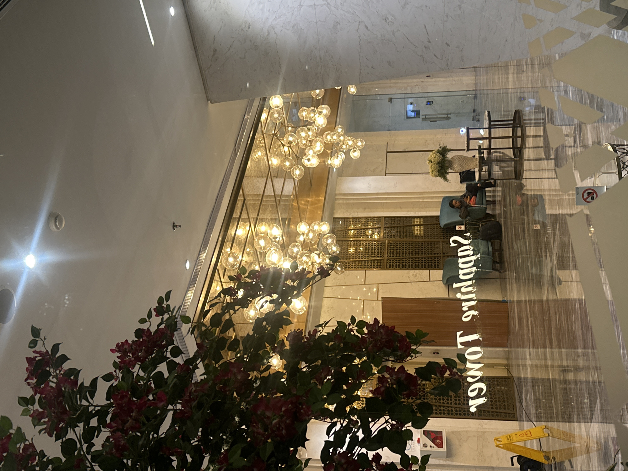The image depicts an interior view of the Sapphire Tower lobby in Central Jakarta, Indonesia. A large decorative chandelier hangs from the ceiling, while a floral arrangement is positioned in the foreground. A person is seated in a blue chair in the background. The name of the building, "Sapphire Tower," is displayed in reflective lettering on a glass surface.