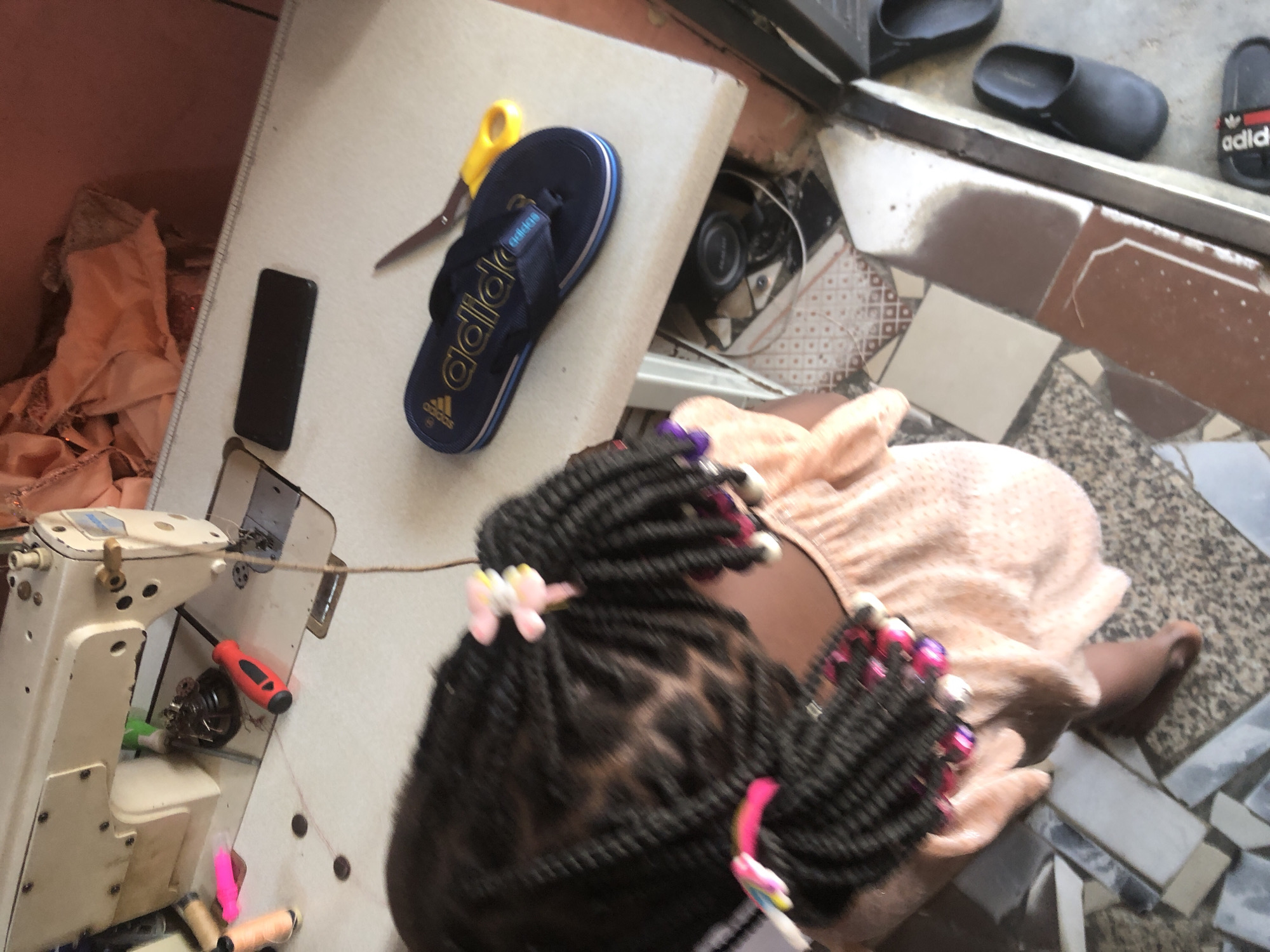 The image depicts a sewing machine set up on a table. A young girl with braided hair, adorned with colorful beads and hair clips, is seated next to the table, her back partially towards the camera. She is wearing a light-colored, ruffled top. On the table, in addition to the sewing machine, are various sewing supplies, including spools of thread, a red and black screwdriver, a pair of yellow-handled scissors, and a dark blue flip-flop. A black smartphone is also visible on the table. The flooring appears to be tiled. The overall setting suggests a domestic or small workshop environment, likely in Ado, Nigeria, given the context provided.