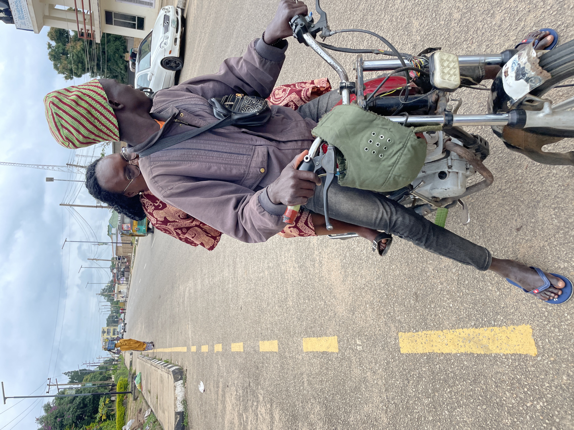 In Jos, Nigeria, a man in a brown jacket and jeans, wearing a green and red striped hat, rides a motorcycle with a woman behind him. The woman wears a red patterned dress. They are on a paved road with yellow dashed lines marking lanes, typical of Jos city. A white car is visible in the background, along with buildings and streetlights. A woman carrying a load on her head walks on the sidewalk in the distance. The scene suggests ordinary urban life in Jos, possibly a busy street.