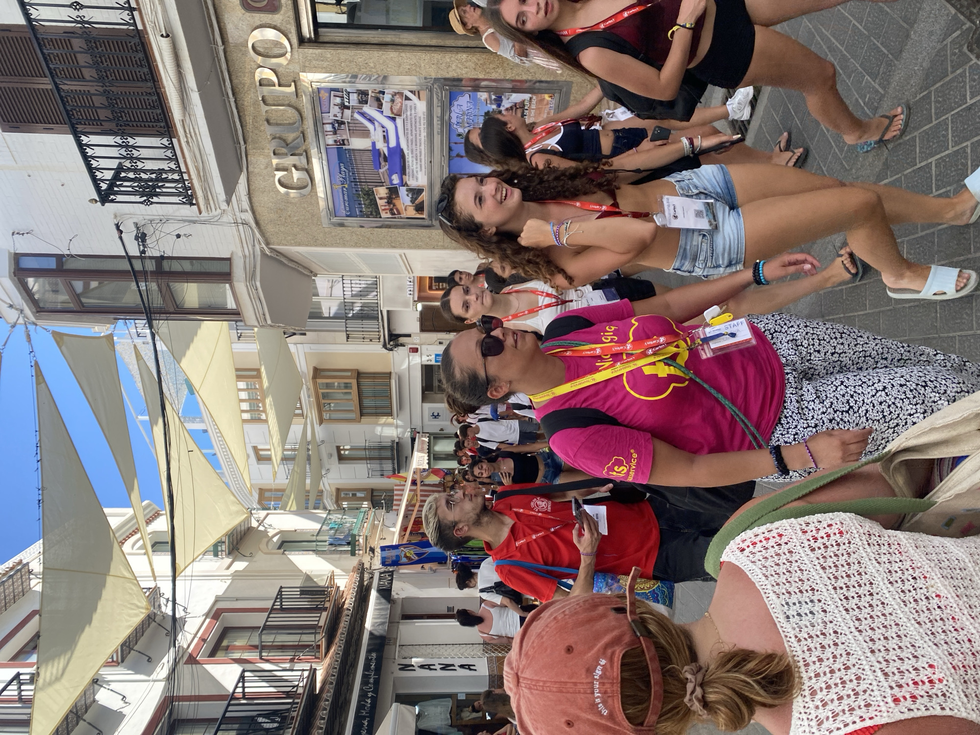 The image depicts a sun-drenched street in Nerja, Spain, characterized by its narrow buildings adorned with balconies and awnings that cast shadows. A group of people, appearing to be tourists or attendees of an event, are walking down the cobblestone street. Many are wearing lanyards with identification badges, suggesting a planned gathering.
In the foreground, a person wearing a red baseball cap with the inscription "this is your sign" is partially visible, facing away from the camera. To their right, a woman in a bright pink t-shirt with a yellow graphic and "its" logo is walking, also wearing a lanyard. She is followed by a young woman with curly brown hair, wearing a dark bikini top and denim shorts, laughing and looking to her left. Another woman, wearing a dark red tank top and black shorts, is walking to the right of the frame.
The background shows more people walking, shops with signage like "NANA," and buildings with traditional Spanish architecture, including shuttered windows and wrought-iron railings. The sky is clear and blue. The overall scene conveys a lively atmosphere during daytime, likely a warm summer day in a popular tourist destination. The presence of the canvas awnings suggests a deliberate effort to provide shade for pedestrians in the bustling street.