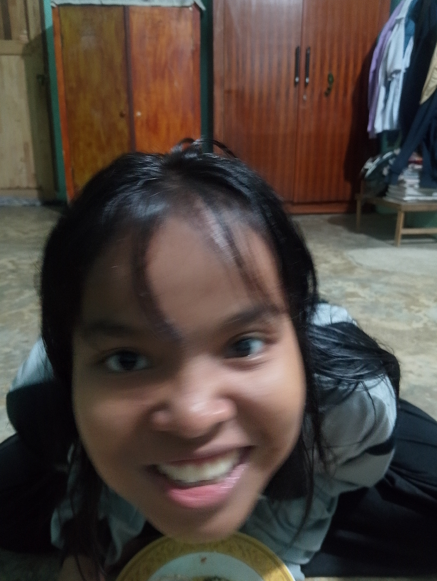 In Serbelawan, Indonesia, a close-up, low-angle shot captures a young person's face, filling most of the frame. Their dark hair frames a smiling face, with teeth visible. The person is wearing a gray top with a black collar and dark pants. Behind them, a wooden wardrobe with two doors and metal handles is visible. To the right of the wardrobe, a stack of books sits on a wooden table, with clothing hanging on a rod above it. The floor appears to be tiled, and a wooden wall is visible on the far left.