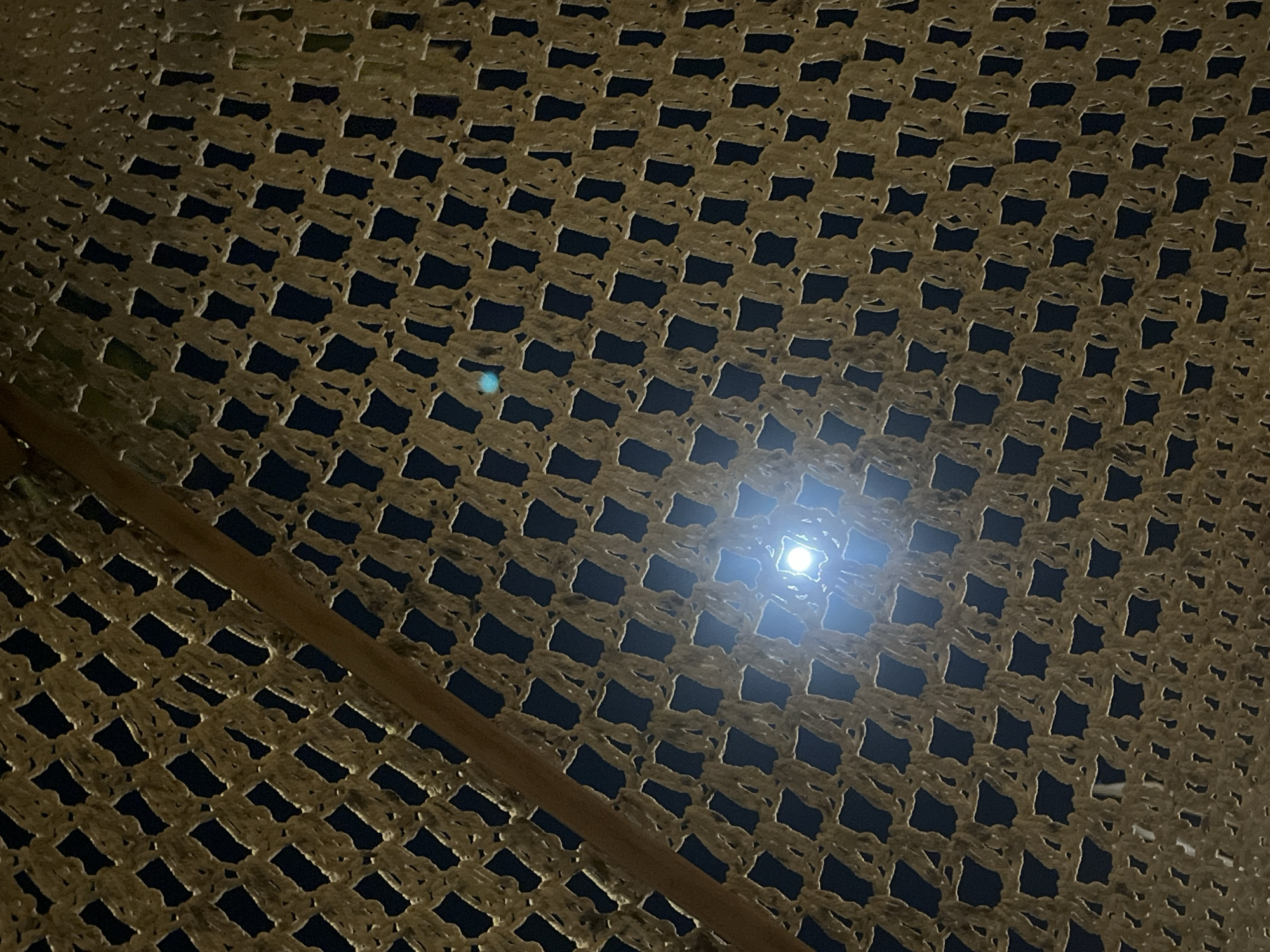 The image, taken at night, appears to be a view upwards through a coarse net or woven material, possibly part of an outdoor structure or shelter. The dark background visible through the netting suggests it is nighttime. A bright light source is visible through the net, creating a focal point in the image. A wooden or bamboo pole is partially visible on the left side of the frame, likely a structural component holding up the net. The lighting and composition suggest a simple, possibly rural, setting. The precise location within Kuta Selatan, Indonesia, cannot be determined from the image alone.