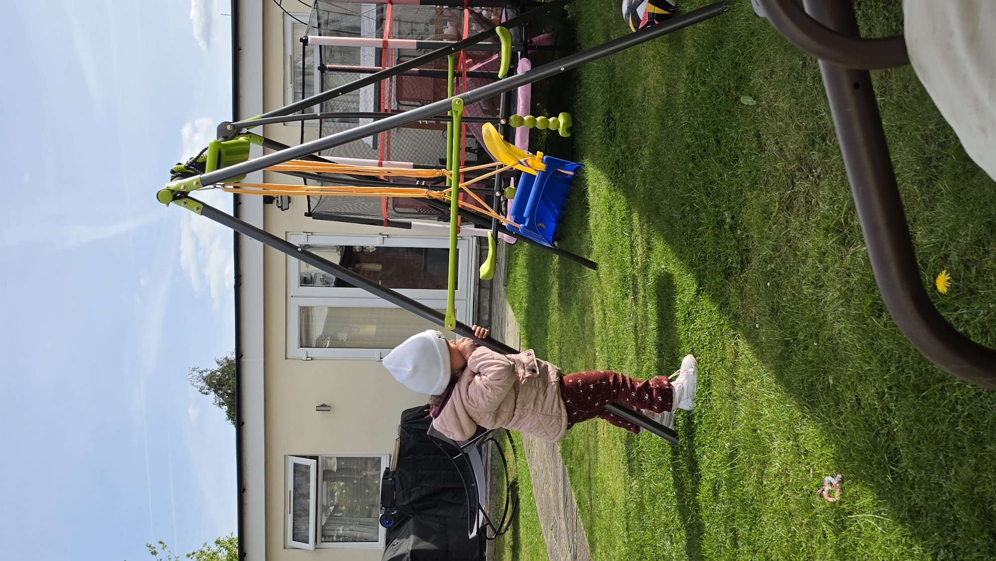 A toddler in a white hat, light pink quilted jacket, and dark red patterned pants is climbing on a black metal swing set in a grassy backyard. The swing set has a green and yellow top bar, orange straps, and a blue swing. In the background is a beige single-story building with white framed windows and a partially visible glass sunroom addition. The sky is blue with light clouds. The location appears to be a residential area, possibly in the United Kingdom, but without further context, a precise location within London cannot be determined.