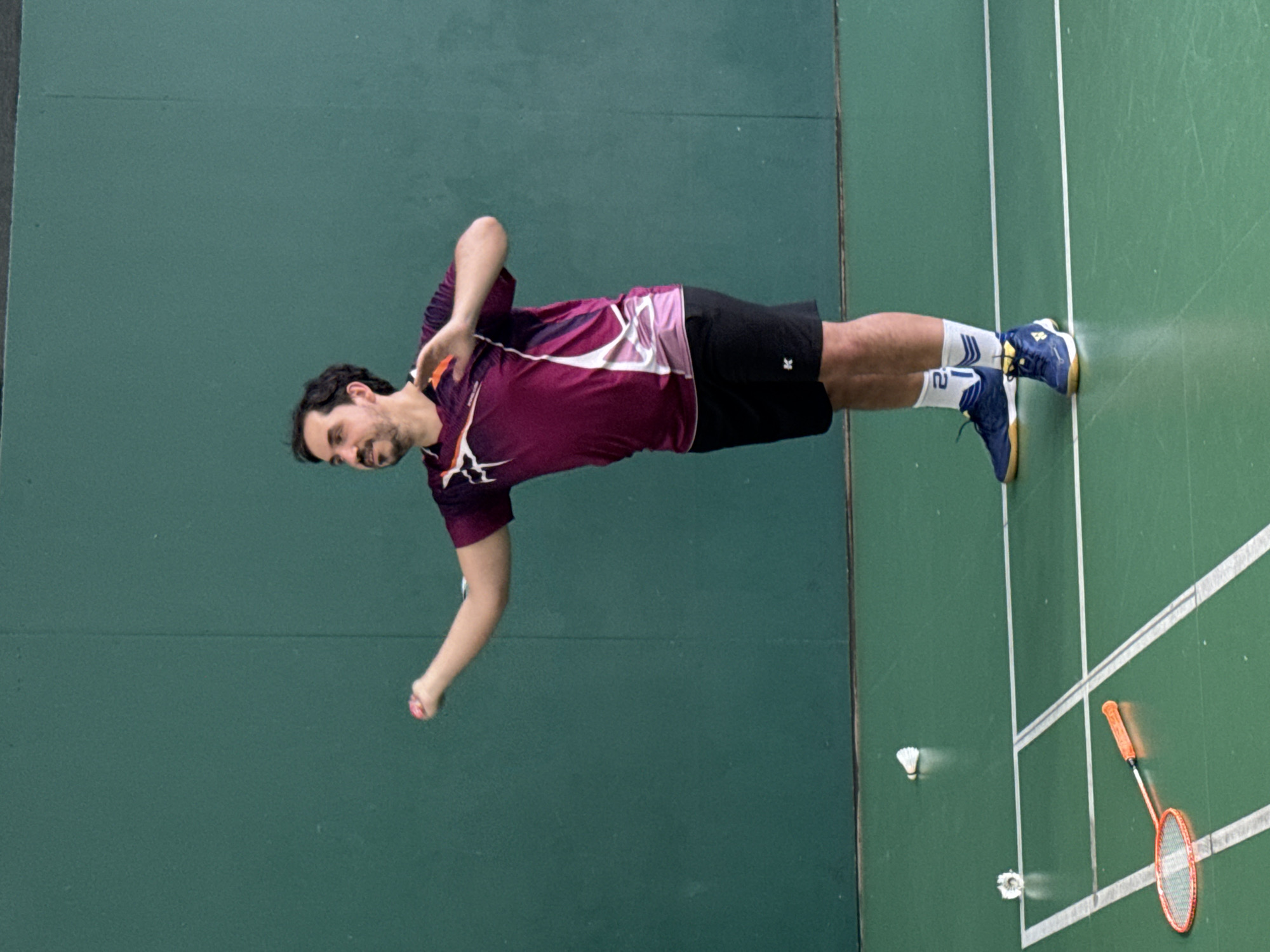 A single adult male is depicted on an indoor green sports court. He is dressed in an athletic maroon and purple short-sleeved shirt with white and orange accents, black athletic shorts displaying a white "K" logo on the left thigh, white socks with blue stripes and the numeral "2" visible, and blue athletic shoes with yellow and white soles. His body is oriented towards the left of the frame, with his left arm extended forward, holding a small red/orange object, likely a badminton shuttlecock. His right arm is bent at the elbow and extended to the right, suggesting a dynamic motion. He has short dark hair and a beard, and his gaze is directed upwards and to the left with a focused expression. On the green court, a red/orange badminton racket and two white shuttlecocks are visible, scattered near the lower left foreground. The court is marked with white lines. The background consists of a solid, dark green wall typical of an indoor sports facility. There is no visual information within the image to confirm the location as the city of Saint Petersburg, Russia.